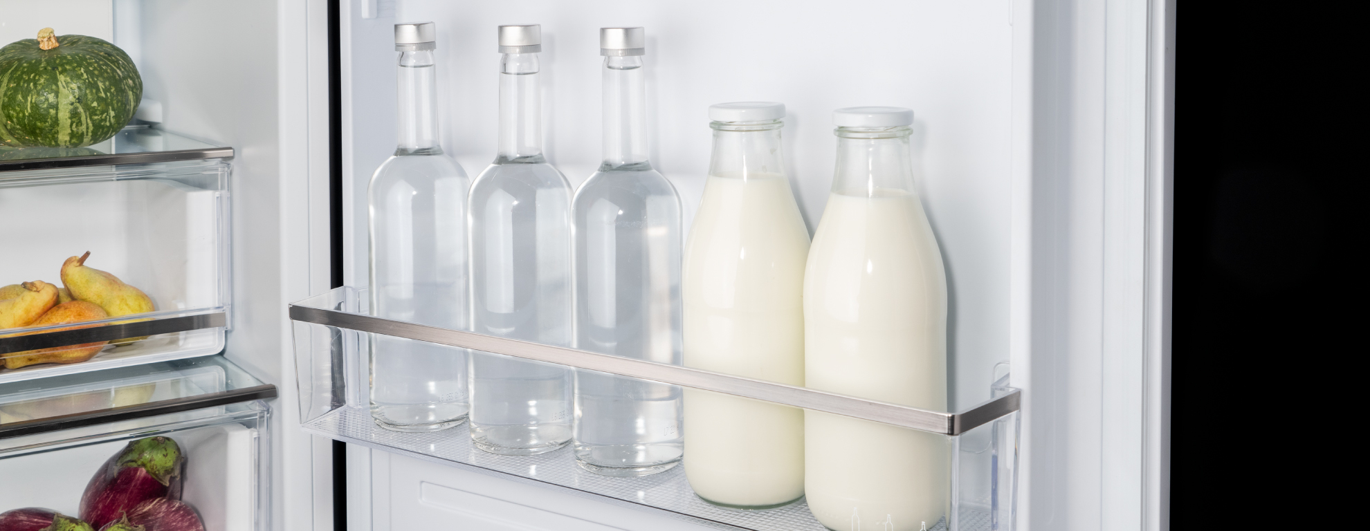 Bottiglie di vetro con acqua e latte nello sportello del frigorifero, frutta sullo sfondo.