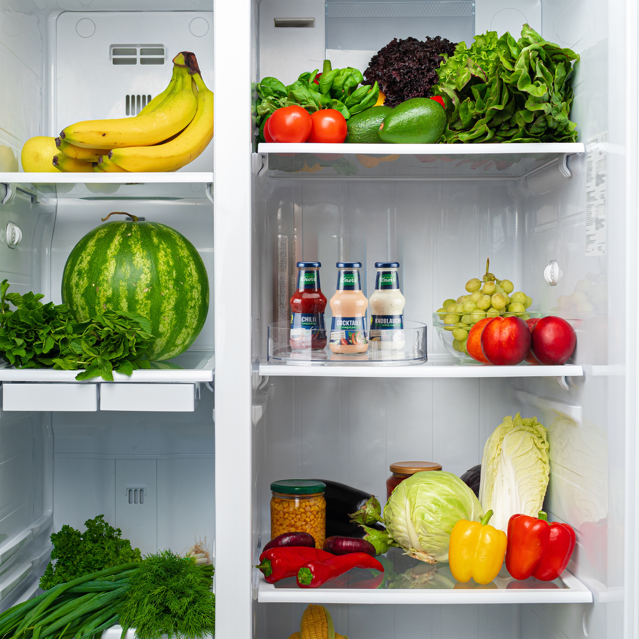 Ein weißer Kühlschrank gefüllt mit frischem Obst, Gemüse, Kräutern und Saucen auf den Regalen.
