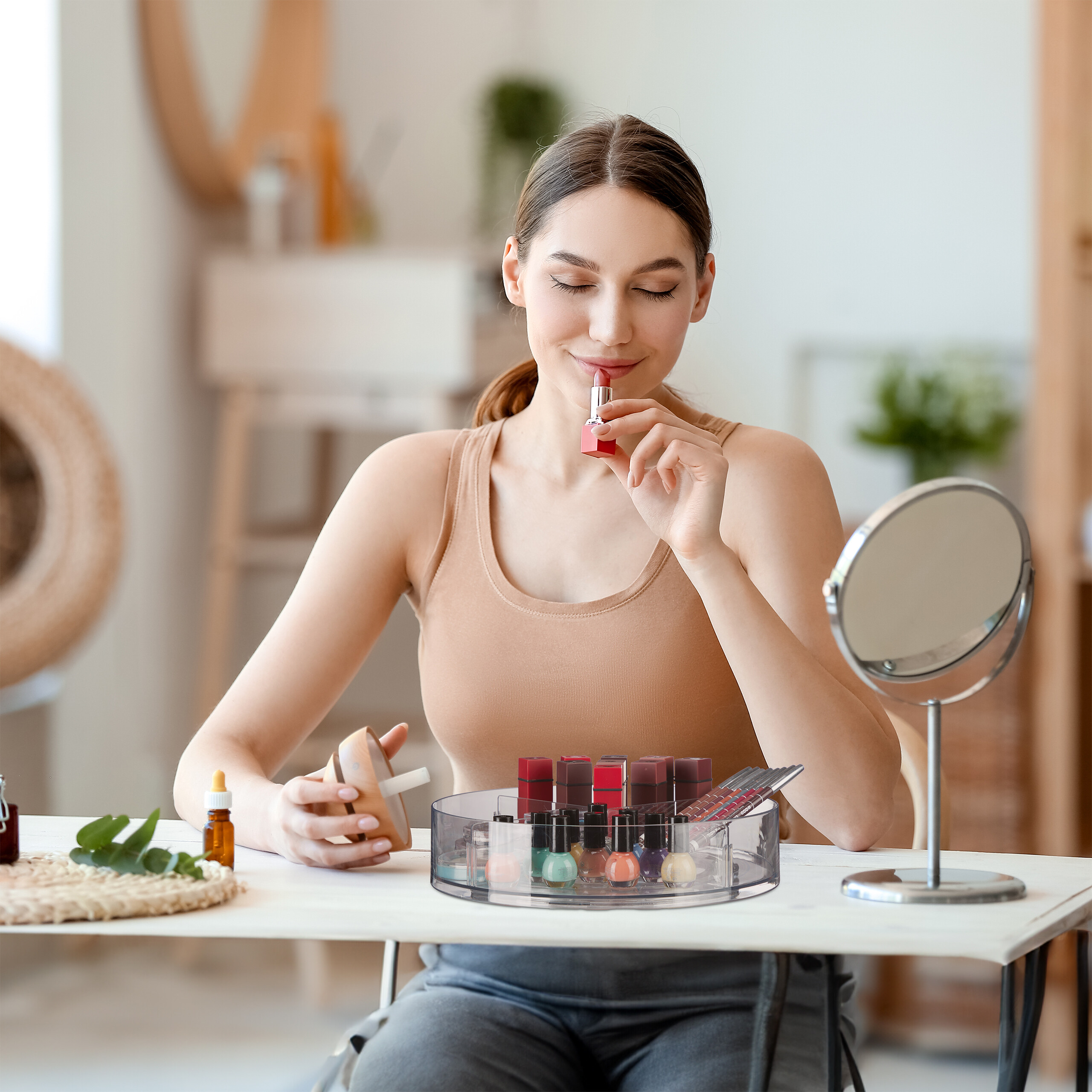 Eine Frau in einem beigen Oberteil sitzt an einem Tisch und hält roten Lippenstift in der Hand, mit Kosmetik davor.