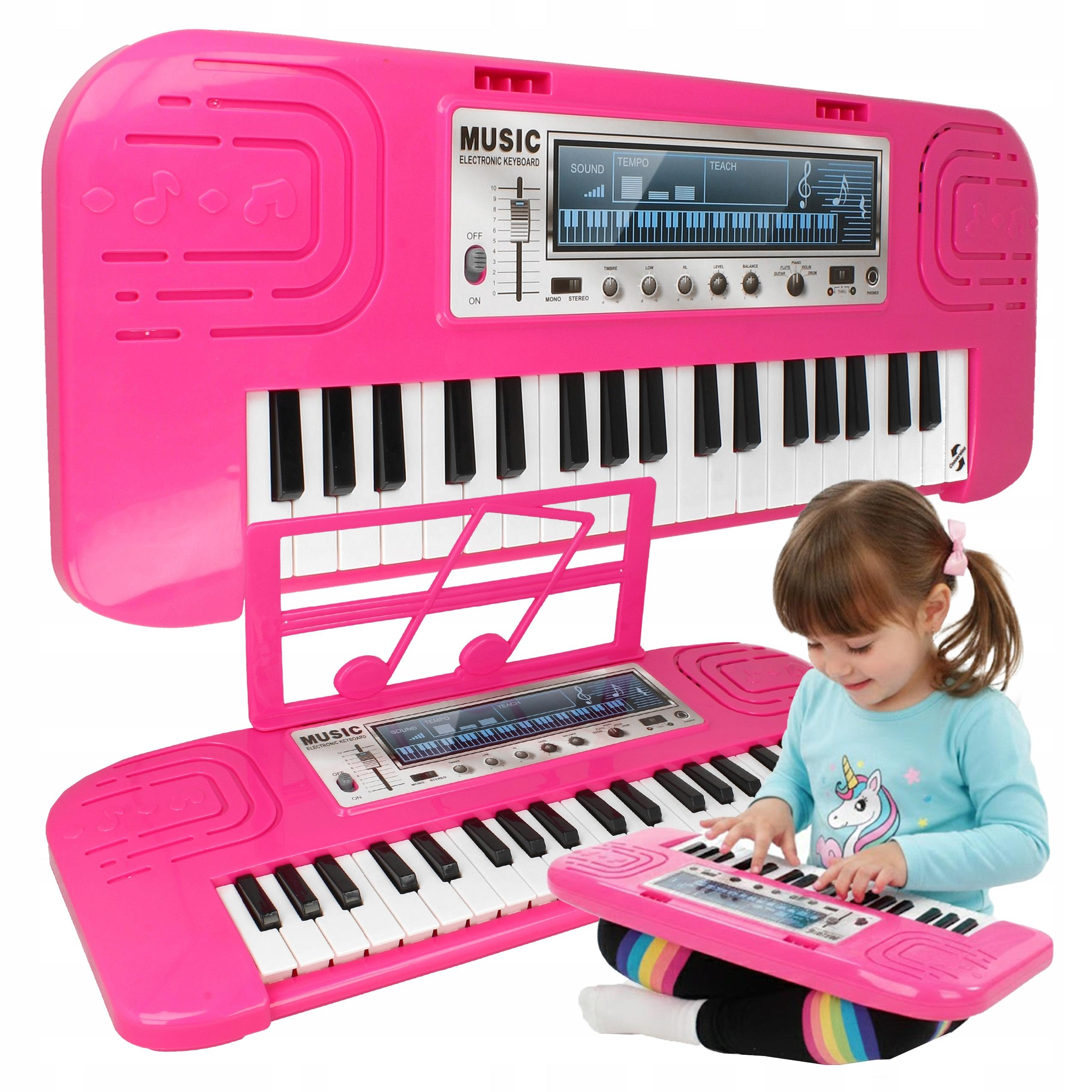 Młoda dziewczynka siedzi i gra na małym różowym keyboardzie zabawkowym, z większymi keyboardami w tle.