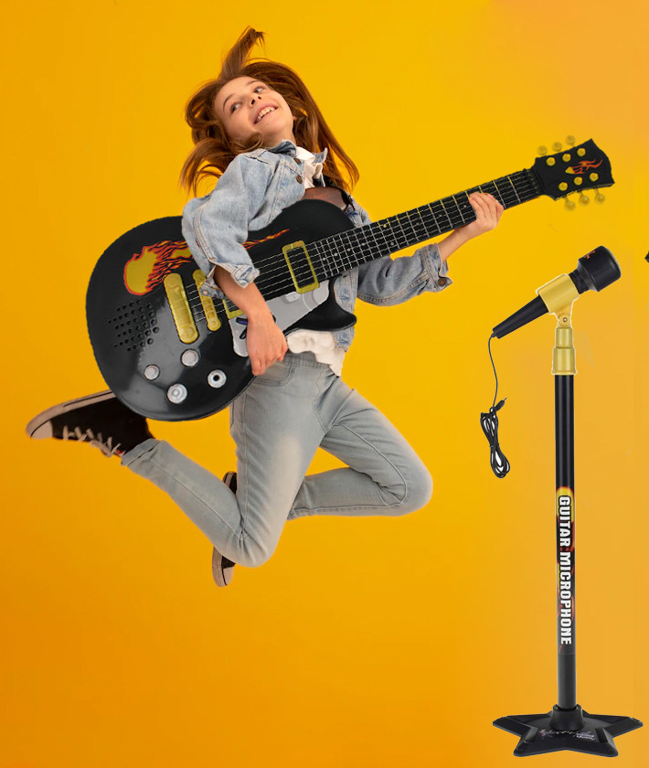 Dziecko w locie gra na czarnej gitarze-zabawce na żółtym tle, obok mikrofonu na statywie.