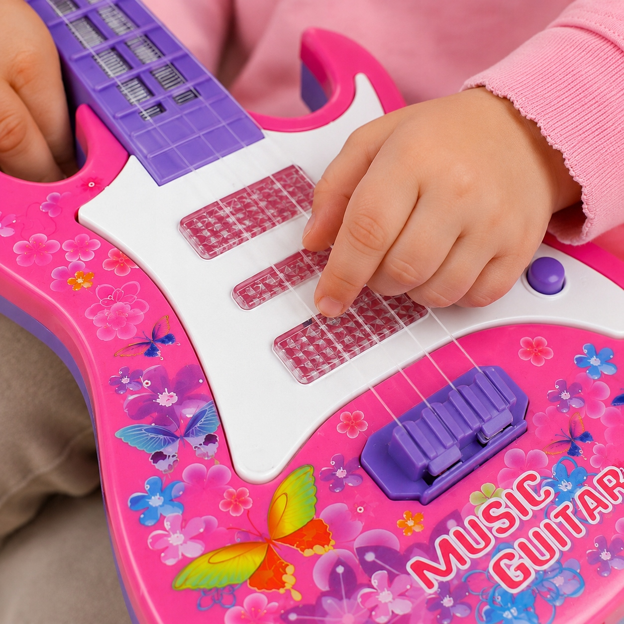 Dziecięce dłonie grają na różowo-fioletowej gitarze-zabawce z motywami kwiatowymi.