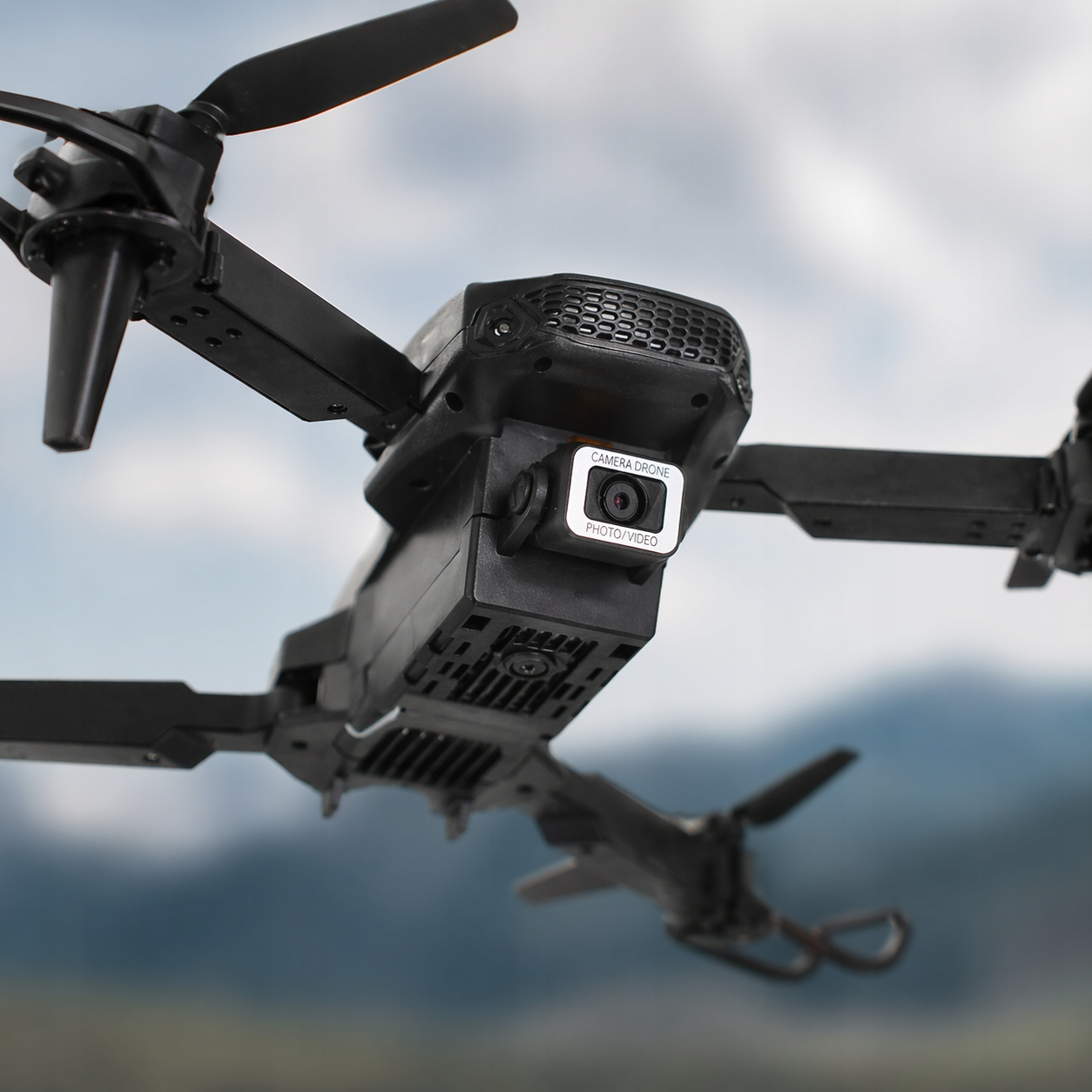 Czarny dron z kamerą unosi się blisko na rozmytym tle pochmurnego nieba i gór.
