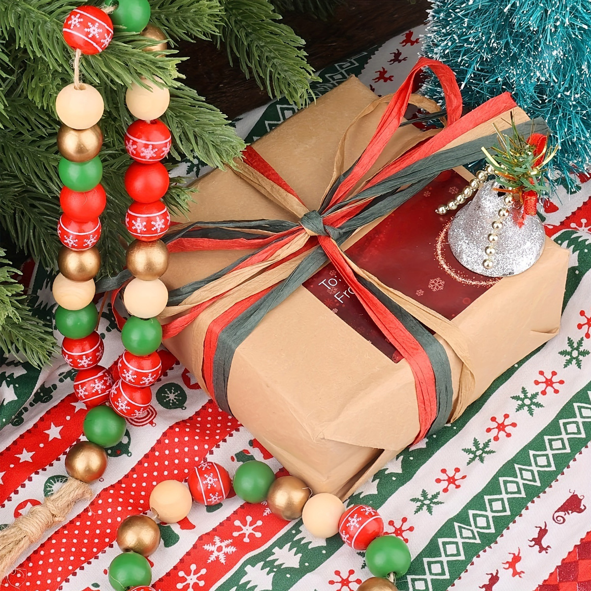 Ein in Packpapier gewickeltes Weihnachtsgeschenk mit roten und grünen Bändern, Perlenketten und Deko auf weihnachtlicher Tischdecke.
