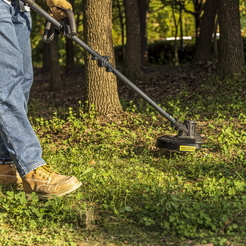 Bir kişi, ağaçlık bir bahçede yabani otları temizlemek için misinalı çim biçme makinesi kullanıyor.
