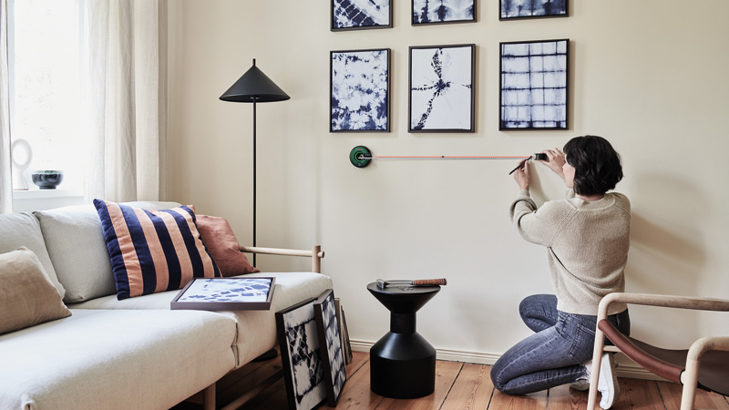 Eine kniende Frau misst mit einem Maßband eine Wand in einem hellen Wohnzimmer mit blauen Mustern an der Wand.