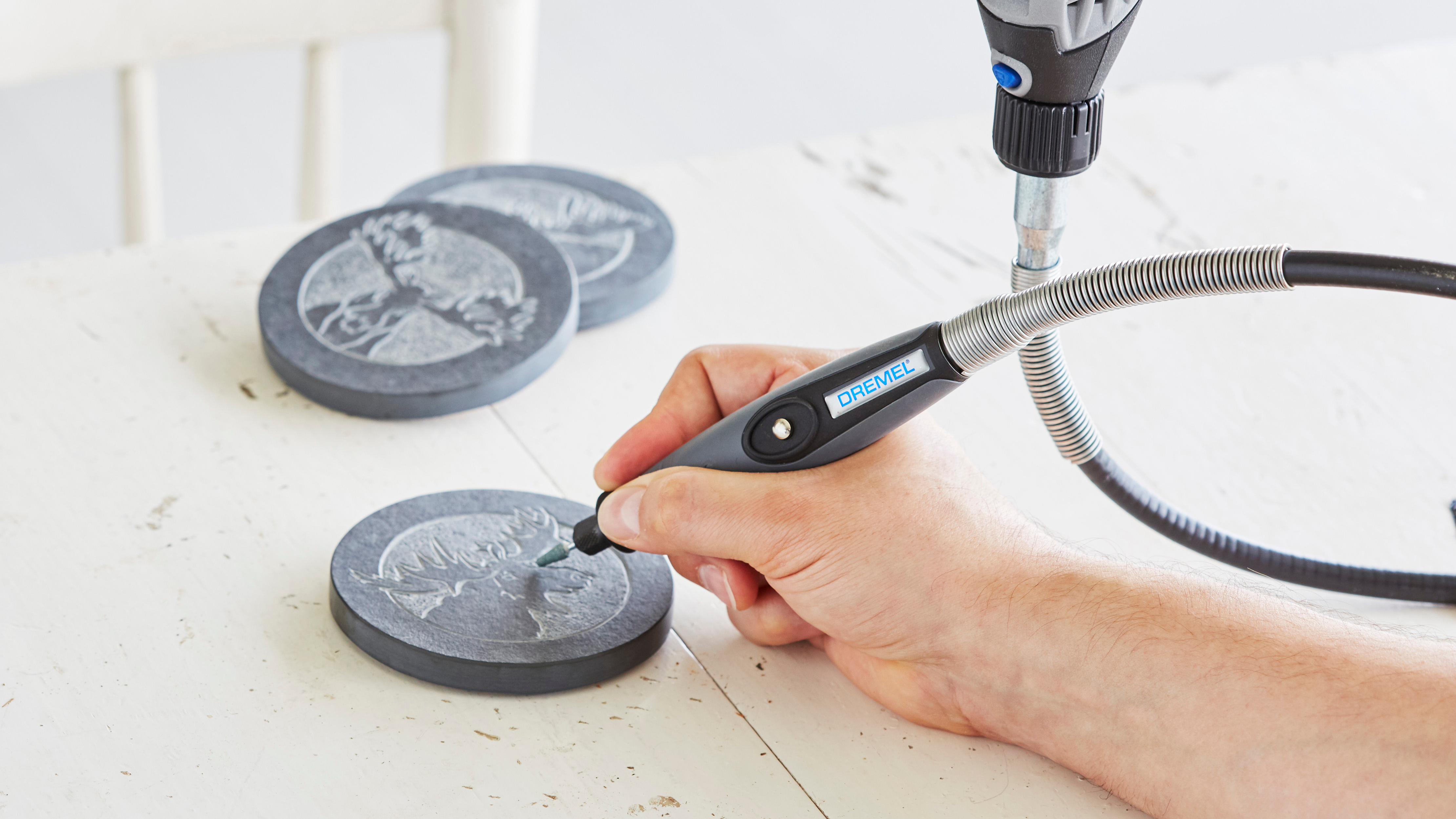 Jemand graviert mit einem Dremel-Fräswerkzeug ein Muster auf einen runden Steinuntersetzer auf einem weißen Tisch.