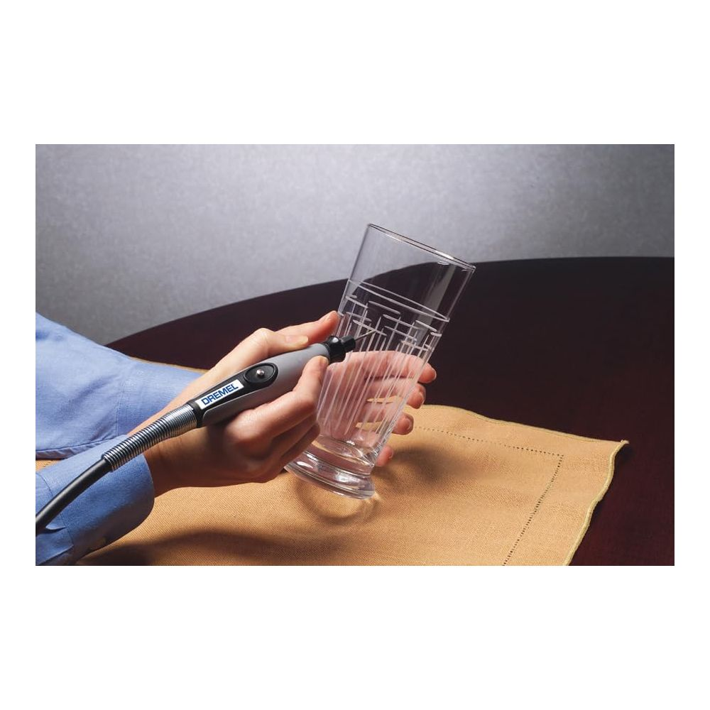 Eine Person graviert mit einem Dremel-Fräser ein geometrisches Muster in ein hohes Glas auf einem Tisch.