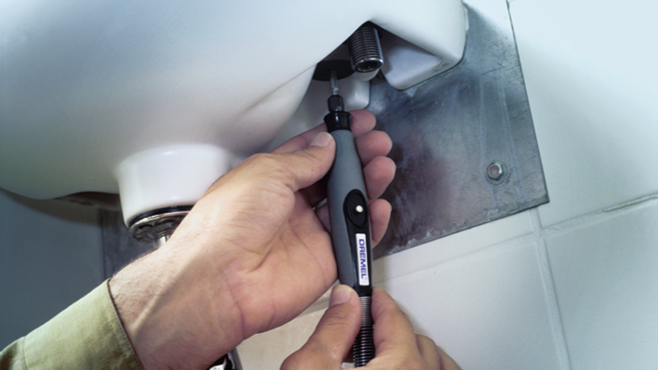 Ein Handwerker repariert mit einem Dremel-Werkzeug die Unterseite eines weißen Waschbeckens an einer gefliesten Wand.