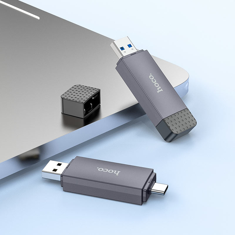 Zwei graue Hoco USB-Sticks und eine Kappe auf heller Oberfläche neben einem Laptop.