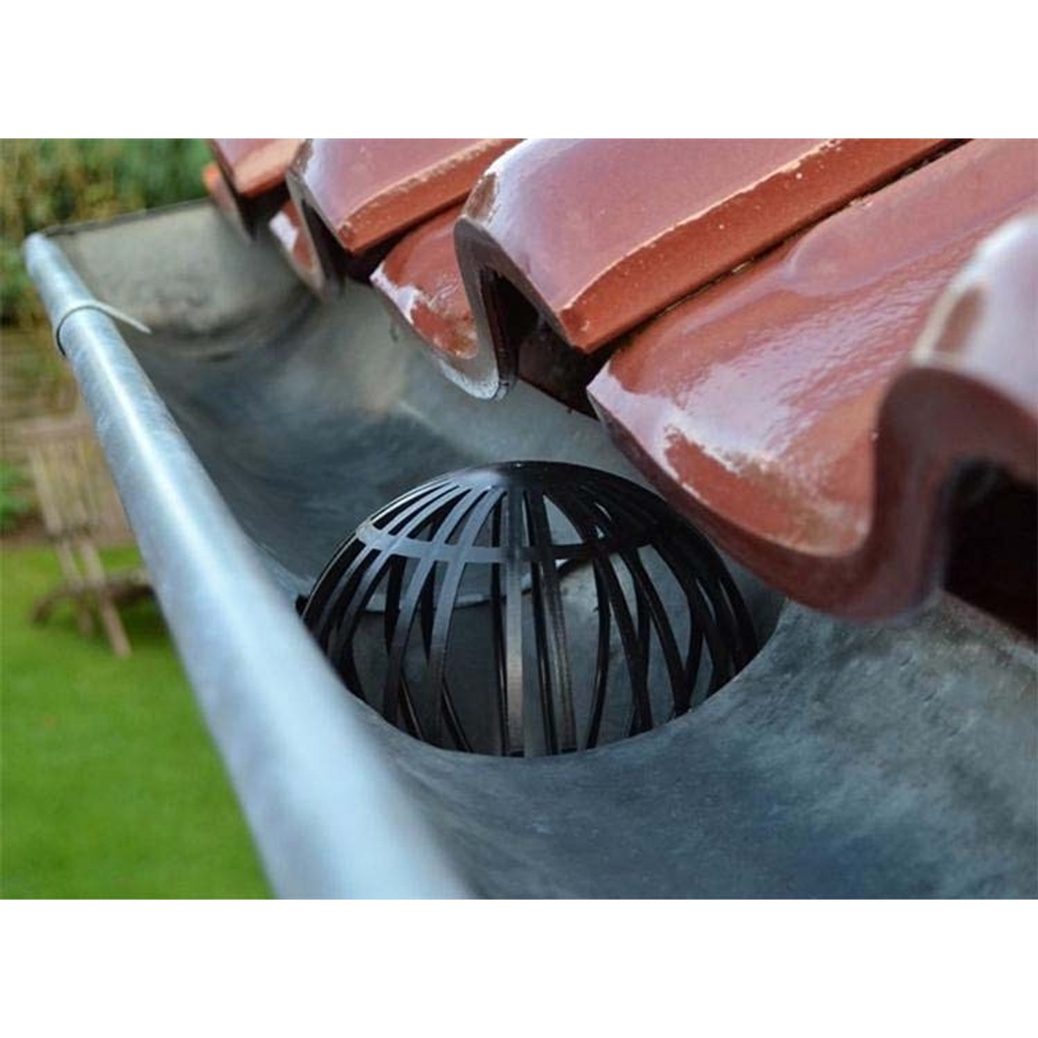 A black spherical cage leaf guard sits in a metallic roof gutter below red roof tiles.