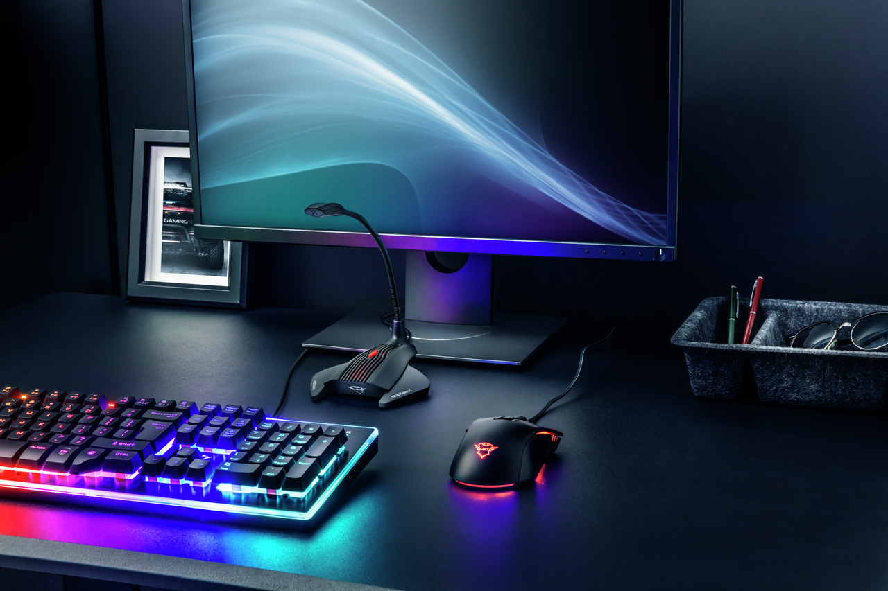 Una postazione da gioco con monitor, tastiera retroilluminata, mouse e microfono in un ambiente scuro.