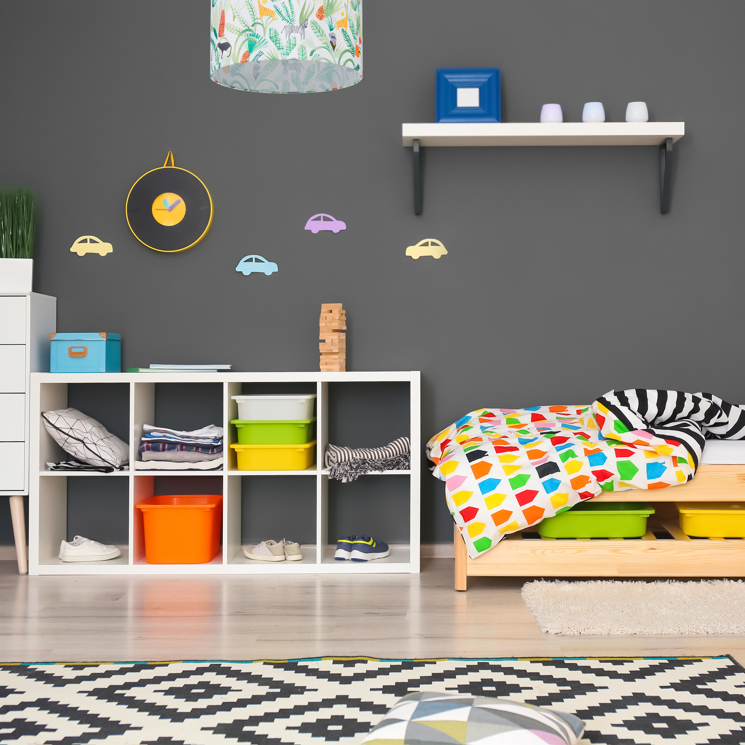 Ein buntes Kinderzimmer mit grauen Wänden, weißem Regal, Bett mit bunter Bettwäsche und gemustertem Teppich.