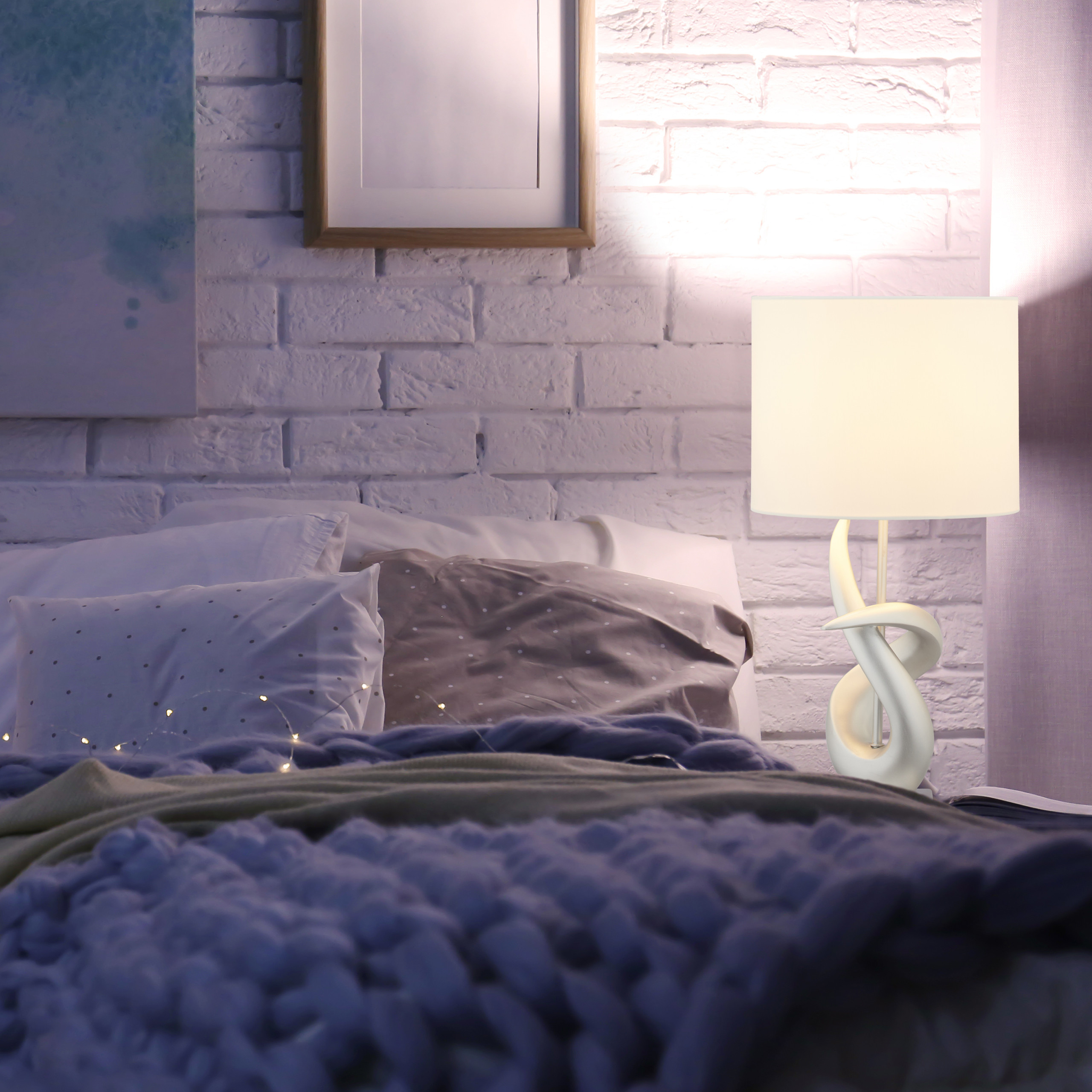 Gemütliches Schlafzimmer mit weißer Backsteinwand, weißer Lampe auf dem Nachttisch und dicker Strickdecke auf dem Bett.