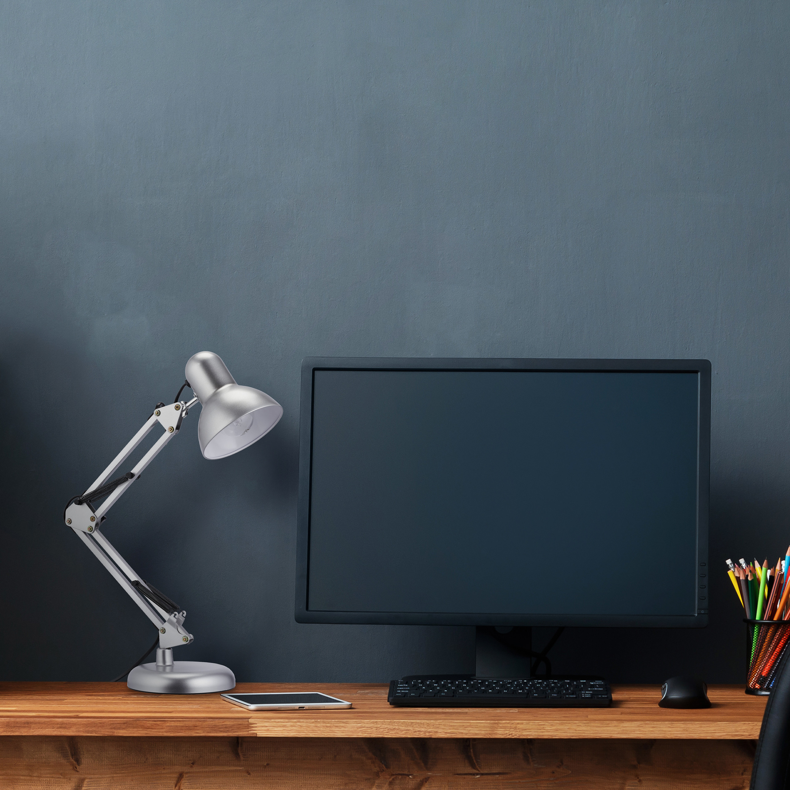Ein Schreibtisch mit Monitor, Lampe, Tastatur und Stiften vor einer blauen Wand.