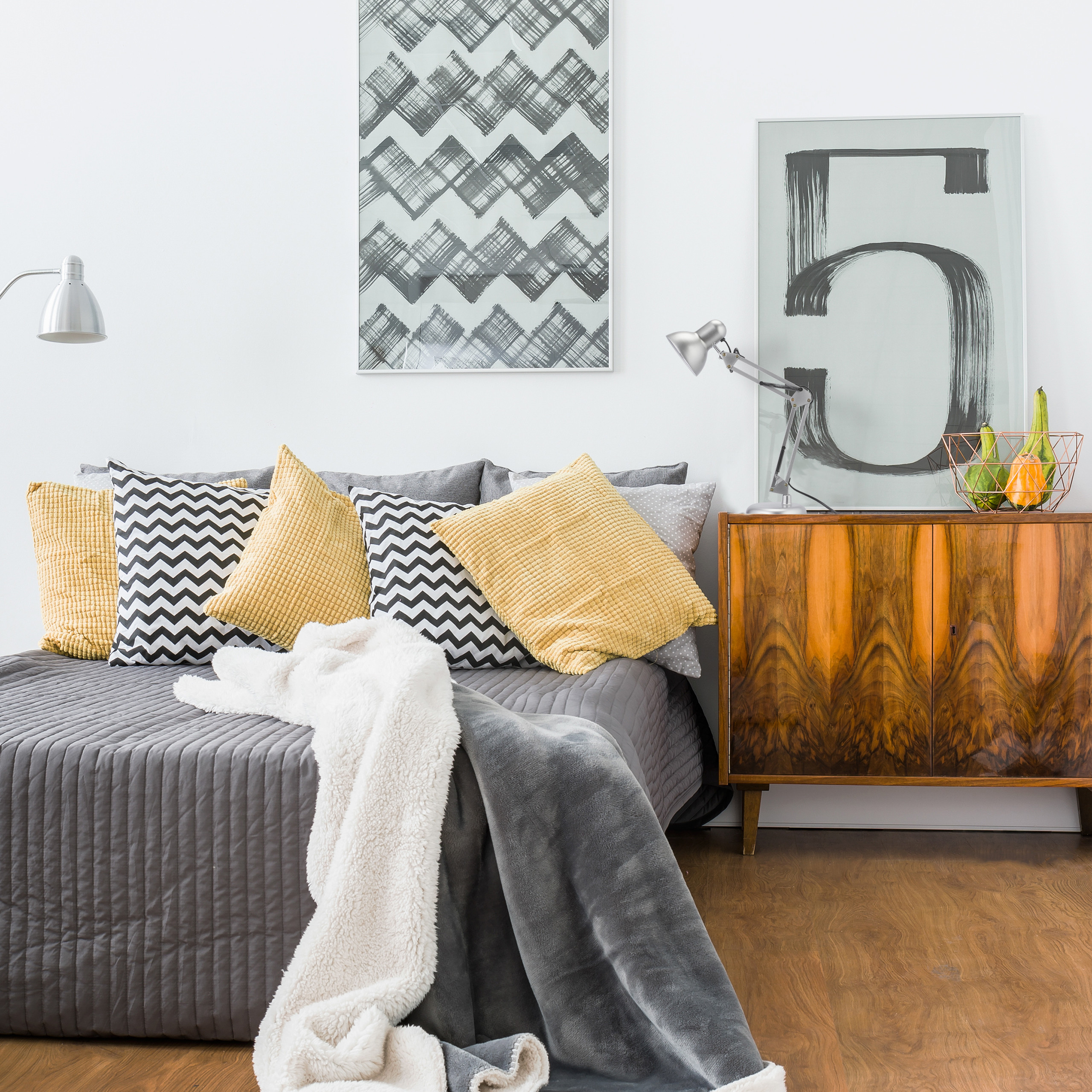 Ein Schlafzimmer mit grauem Steppbett, gelben und gemusterten Kissen, Holzschrank und abstrakter Kunst an weißen Wänden.