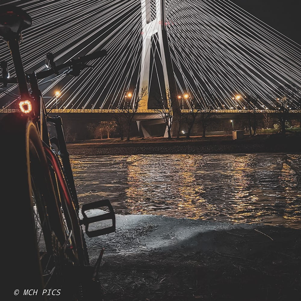 Rower zaparkowany nad rzeką w nocy z oświetlonym mostem wiszącym w tle.