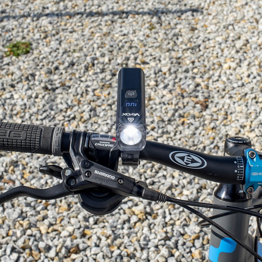 Czarna kierownica roweru z podświetlaną lampką i klamkami hamulcowymi Shimano na tle żwiru.