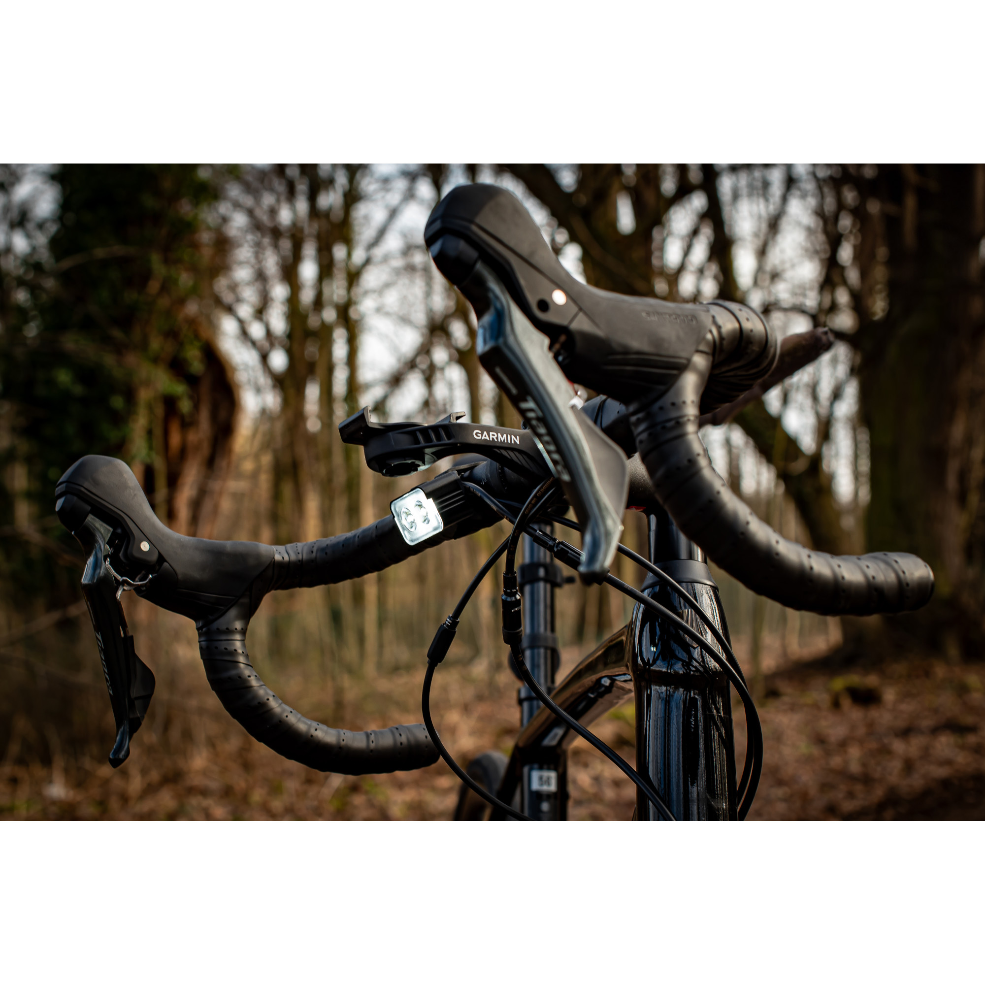 Czarna kierownica roweru szosowego z mocowaniem Garmin i włączoną lampką przednią w lesie.