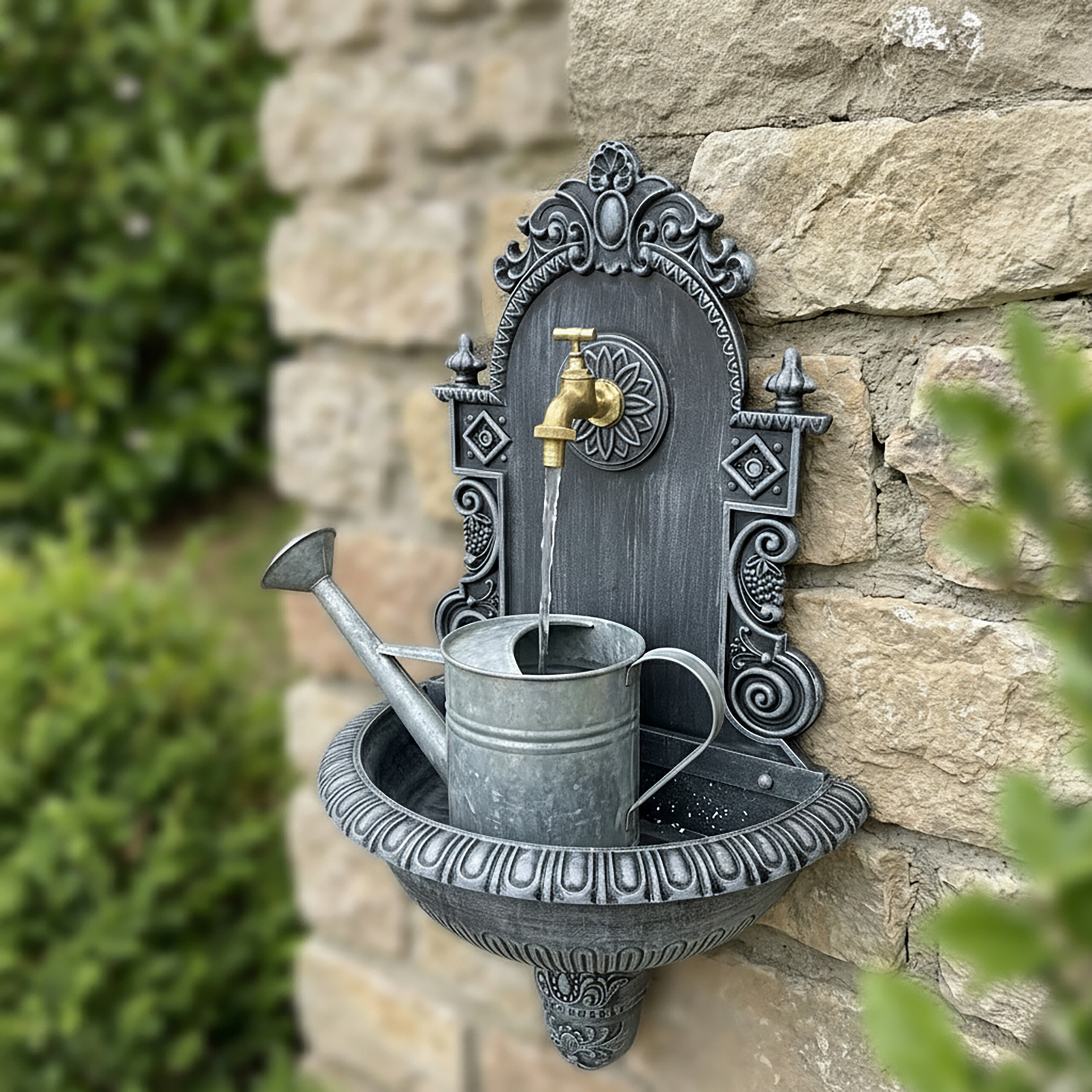 Eine graue, verzierte Wandbrunnenanlage mit goldenem Hahn füllt eine kleine Metallgießkanne vor einer Steinmauer.