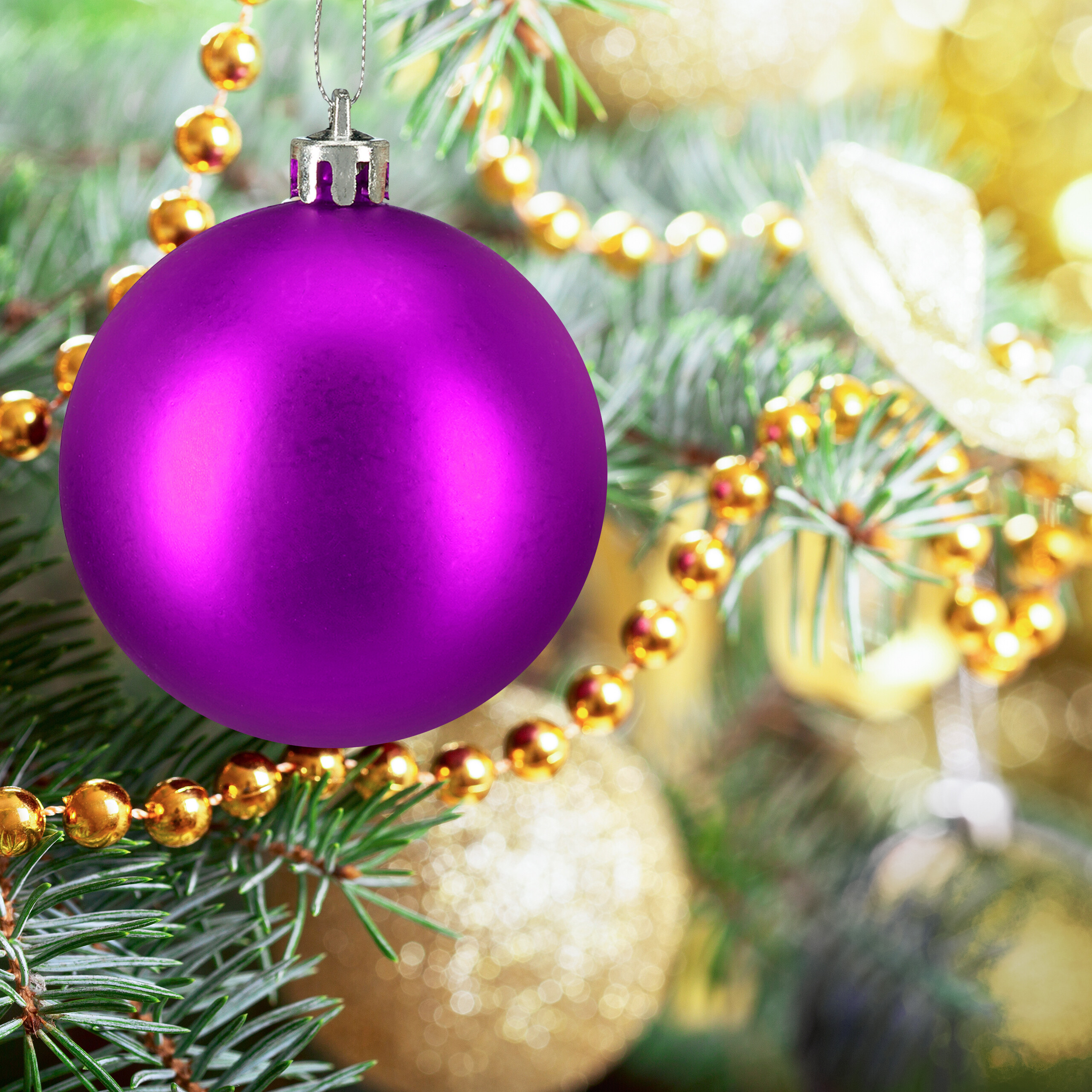 Eine matte, violette Weihnachtskugel hängt an einem grünen Tannenbaum mit goldenen Perlenketten und Lichtern.