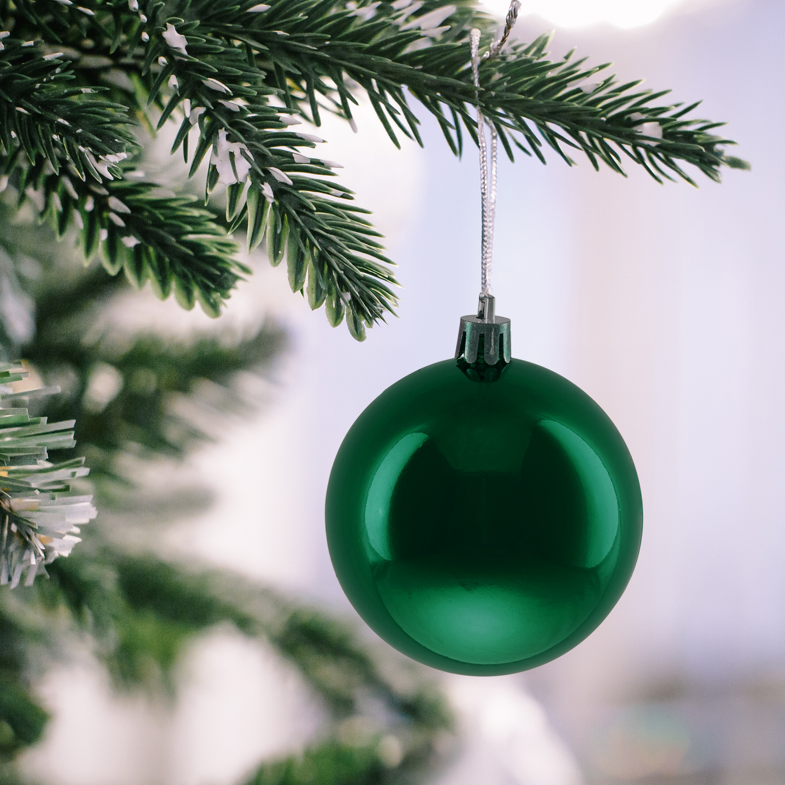 Eine glänzende grüne Weihnachtskugel hängt an einem Tannenzweig mit künstlichem Schnee.