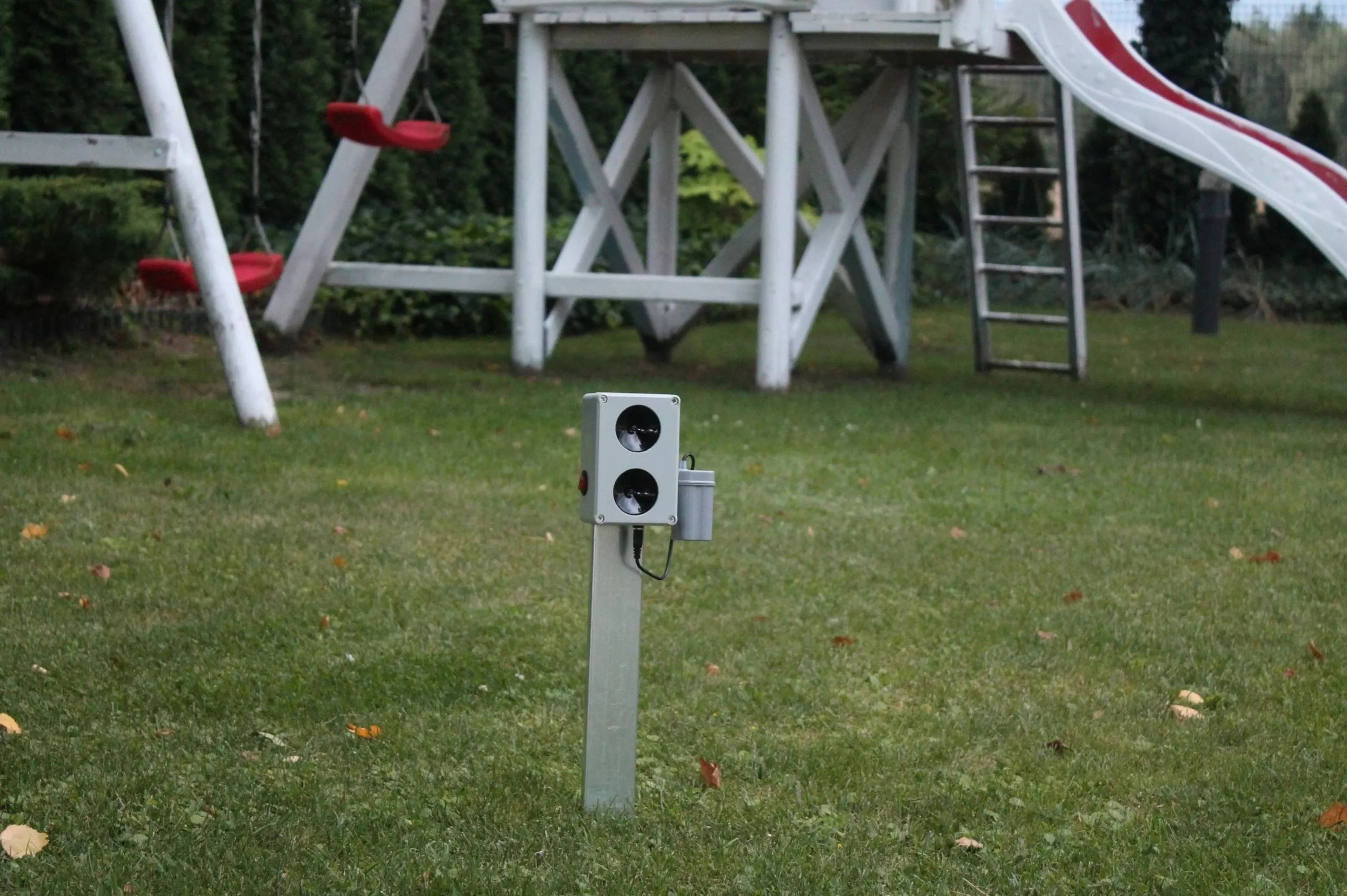 Egy szürke elektronikus eszköz fémoszlopon a kertben, háttérben fehér hintával és csúszdával.