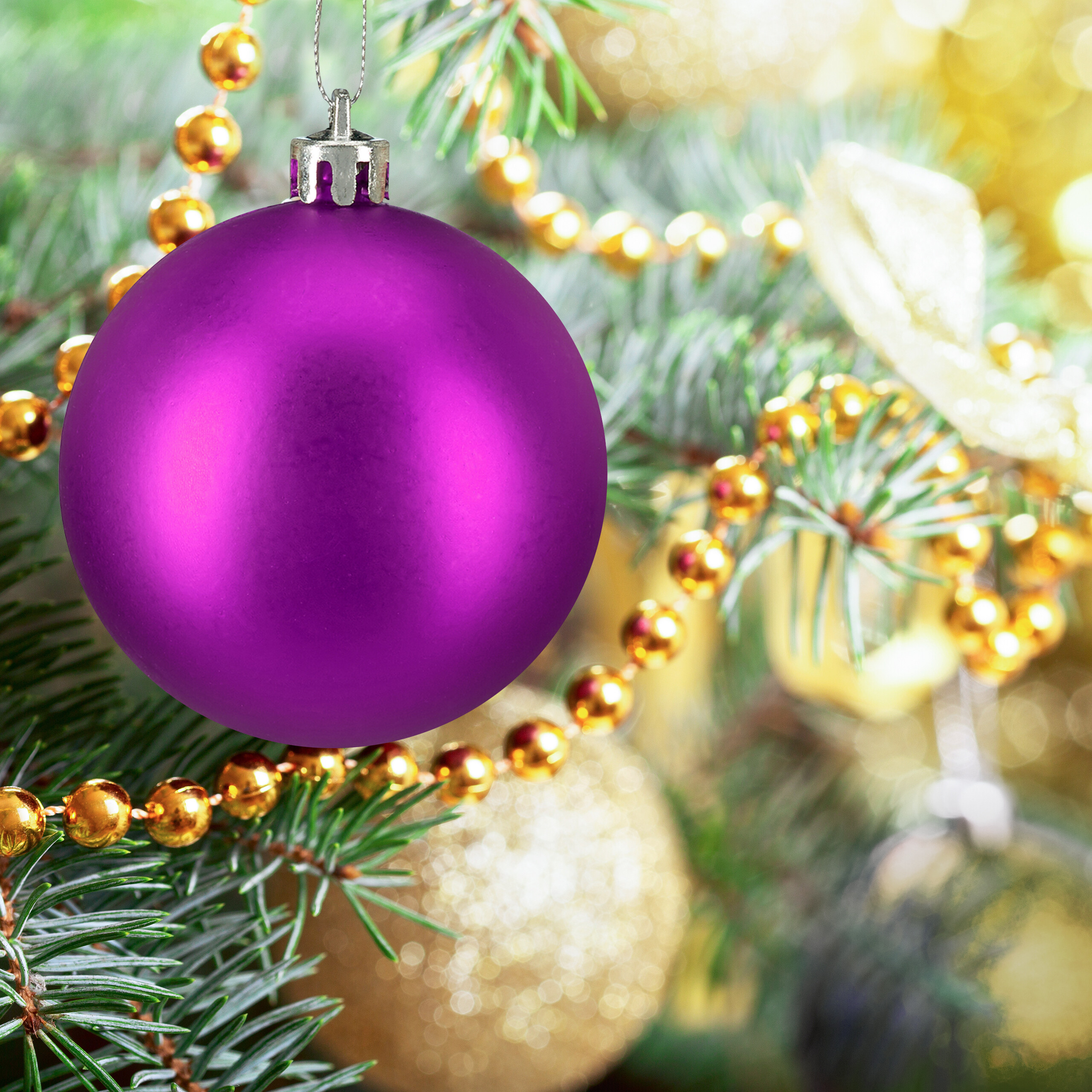 Eine matte, violette Weihnachtskugel hängt an einem geschmückten Tannenbaum mit goldenen Perlen.