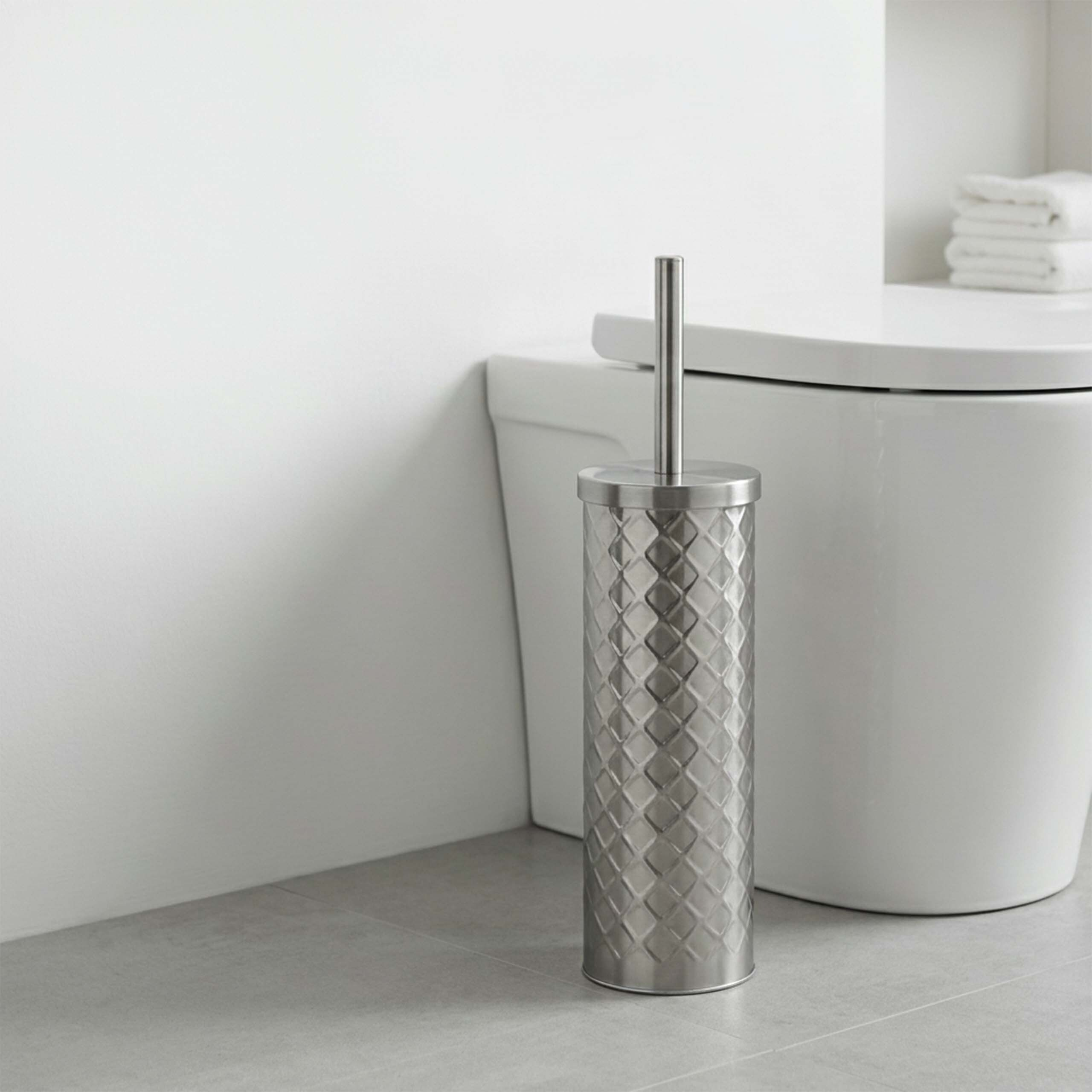 Eine metallische Toilettenbürste mit Rautenmuster steht vor einer weißen Toilette auf dem Boden.
