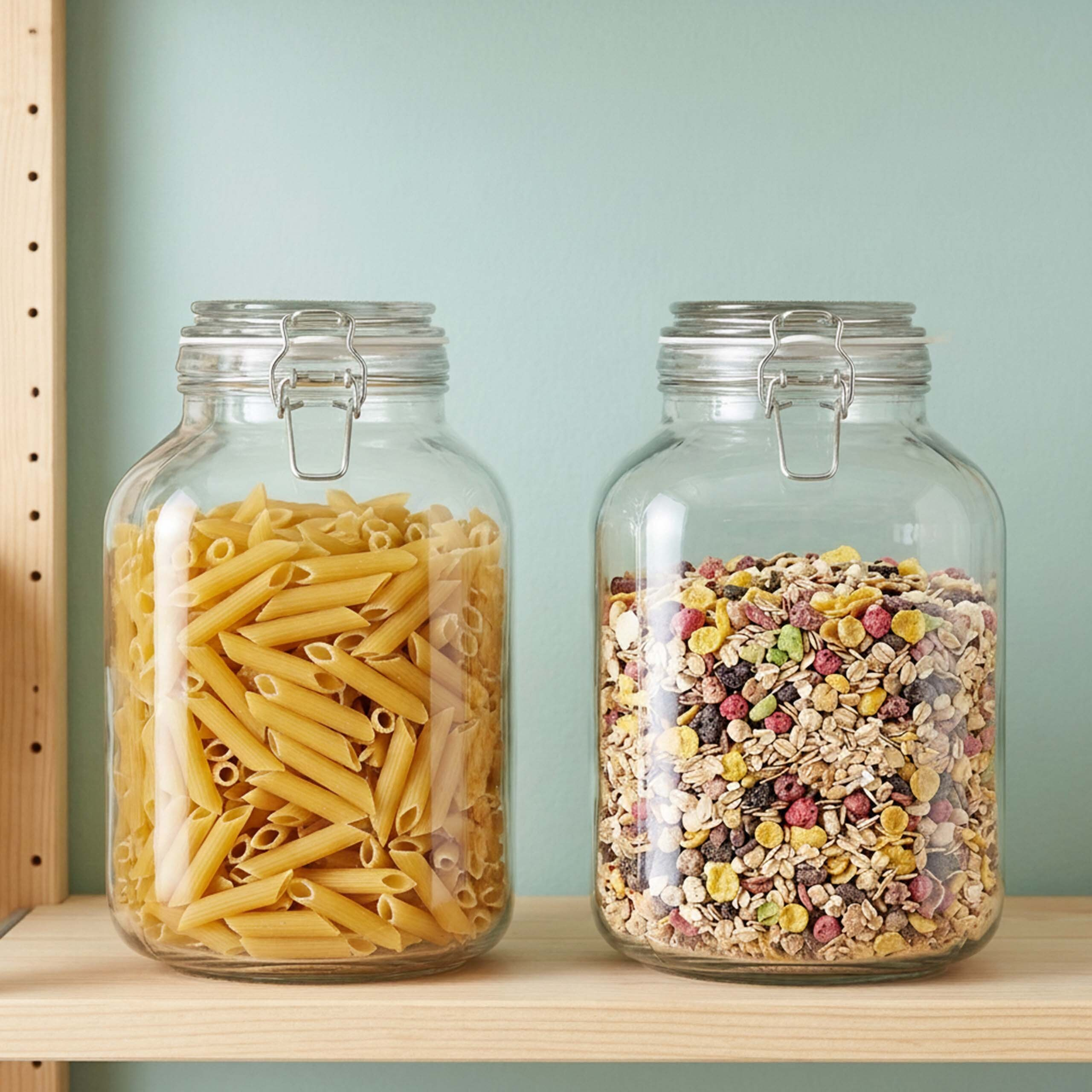 Zwei Glasbehälter mit Metallbügelverschluss auf einem Holzregal vor einer mintgrünen Wand; einer mit Penne, einer mit Müsli.