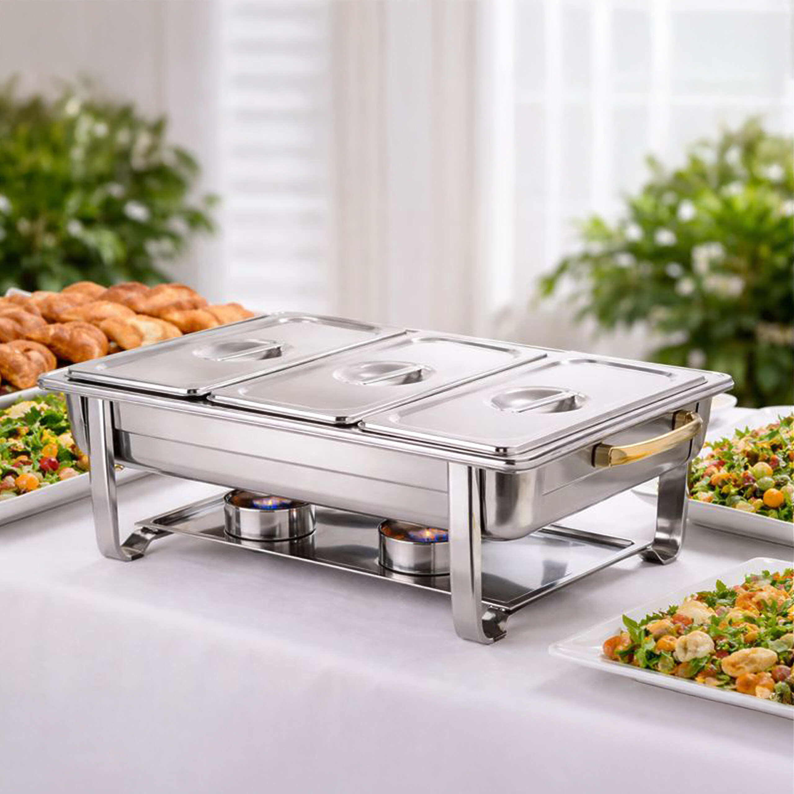 Ein Edelstahl-Chafing-Dish mit drei Deckeln steht auf einem weißen Tisch, umgeben von Schüsseln mit Salat.
