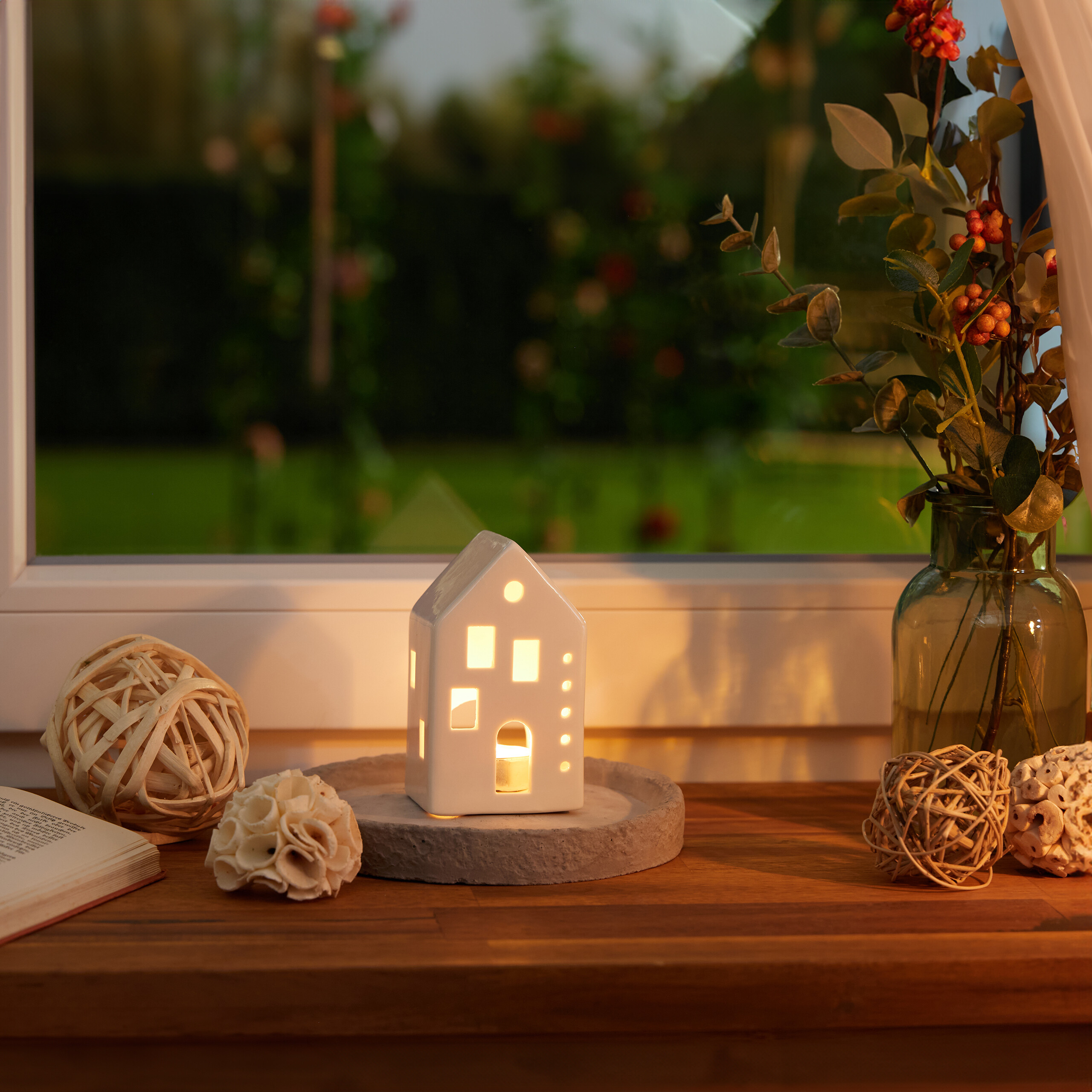 Ein kleines weißes Kerzenhaus auf einem Holzregal mit Blumen, Kugeln und Buch vor einem Fenster mit Gartenblick.