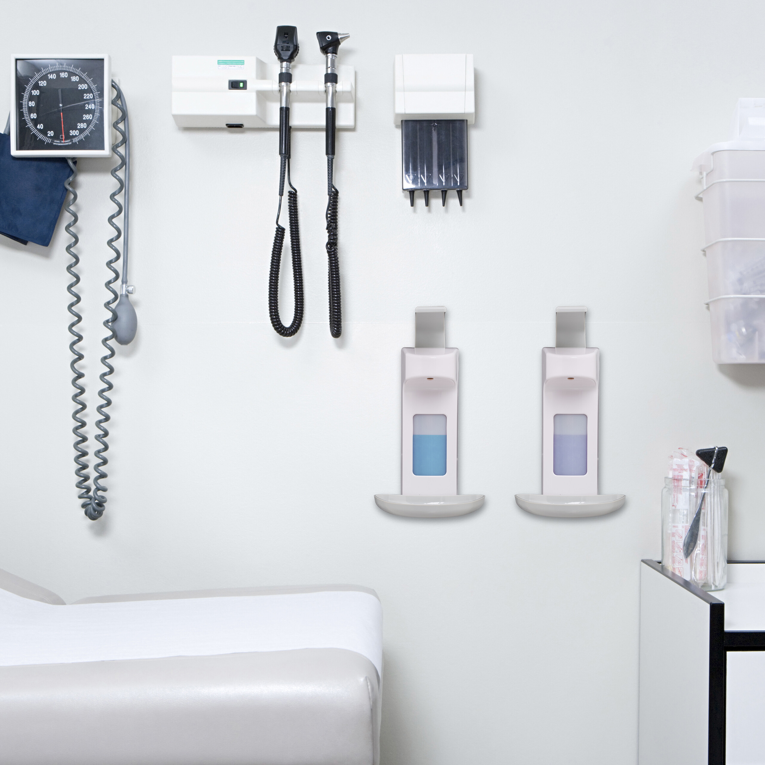 Medizinische Ausrüstung an weißer Wand, darunter Blutdruckmessgerät, Otoskope und Seifenspender in einer Arztpraxis.