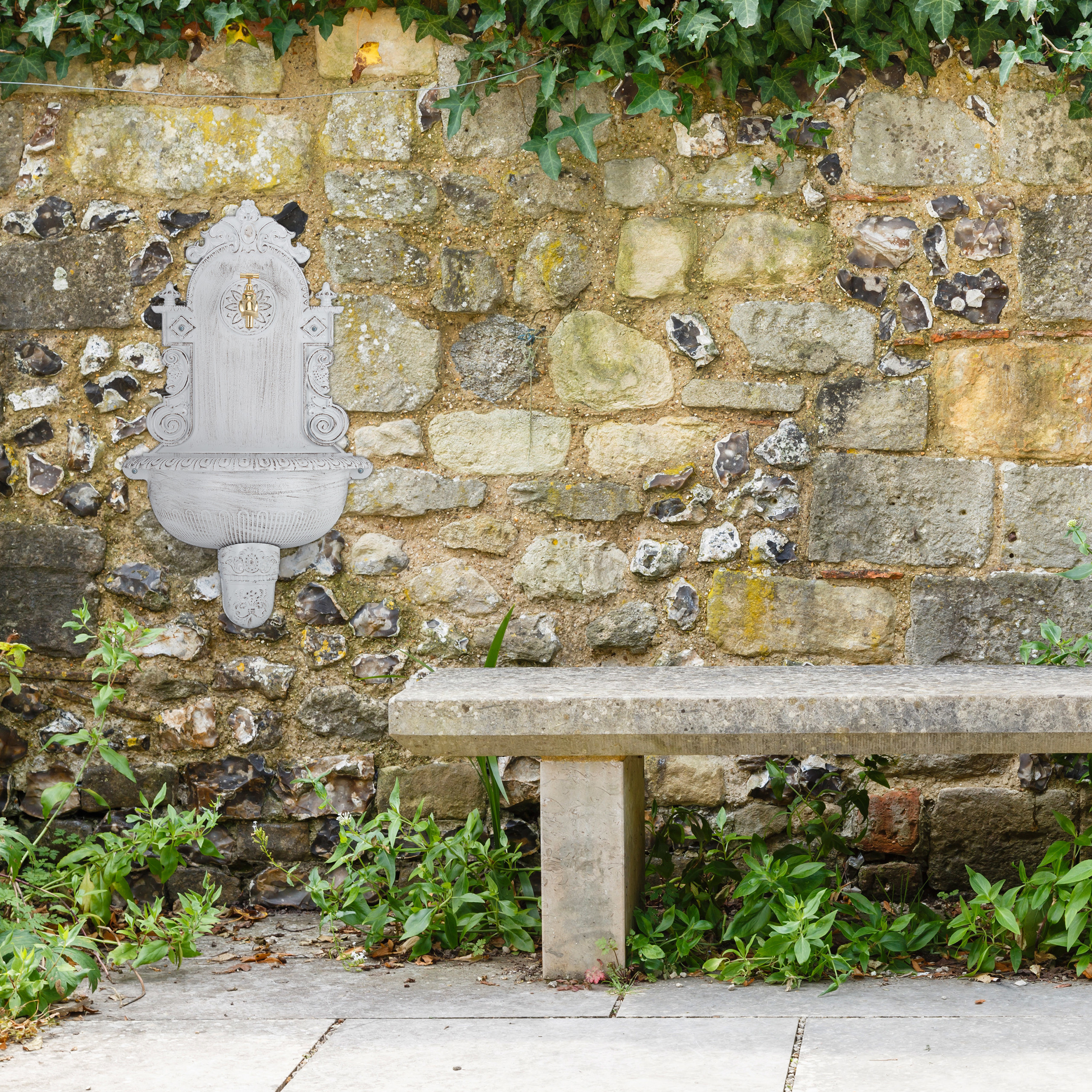 Eine Steinbank steht vor einer verwitterten Steinmauer mit verziertem weißem Wandbrunnen und Efeu.