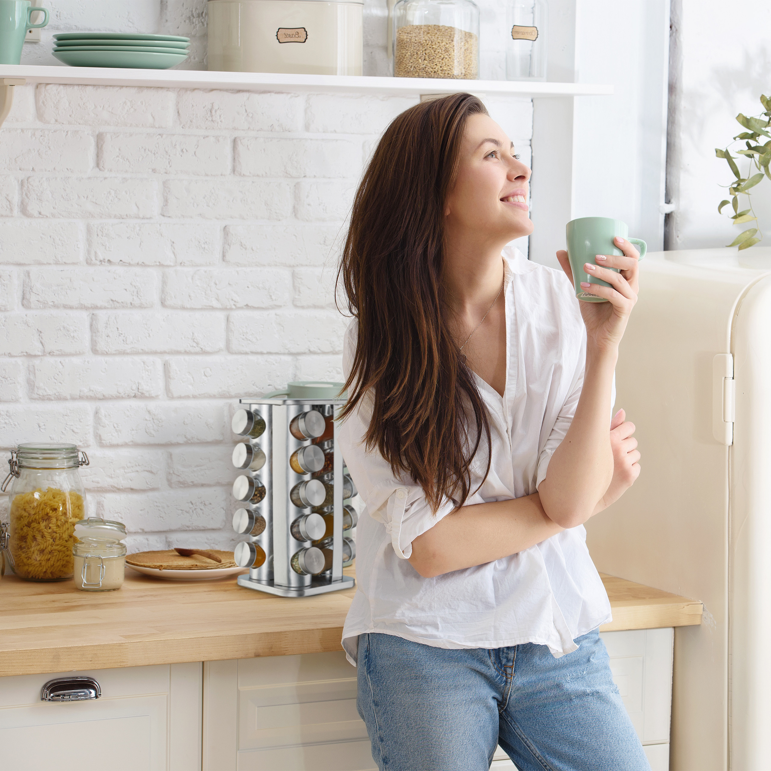 Eine Frau in weißem Hemd und Jeans steht in einer weißen Küche, hält eine grüne Tasse und lächelt.