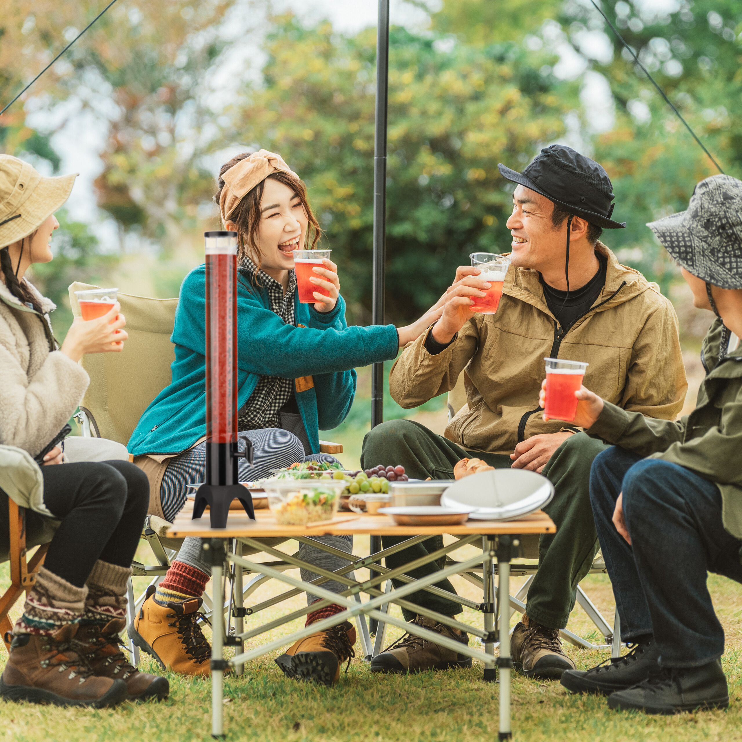 Vier Freunde sitzen beim Camping an einem Tisch, lachen und stoßen mit roten Getränken an.