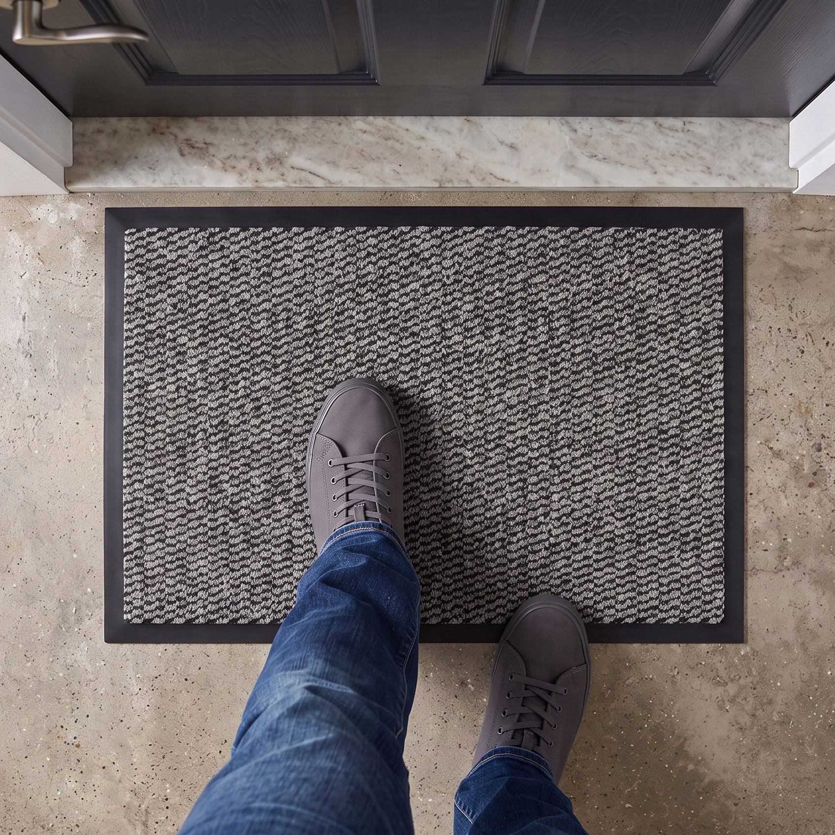 Eine Person steht in blauen Jeans und grauen Sneakers auf einer grauen Fußmatte vor einer Eingangstür.