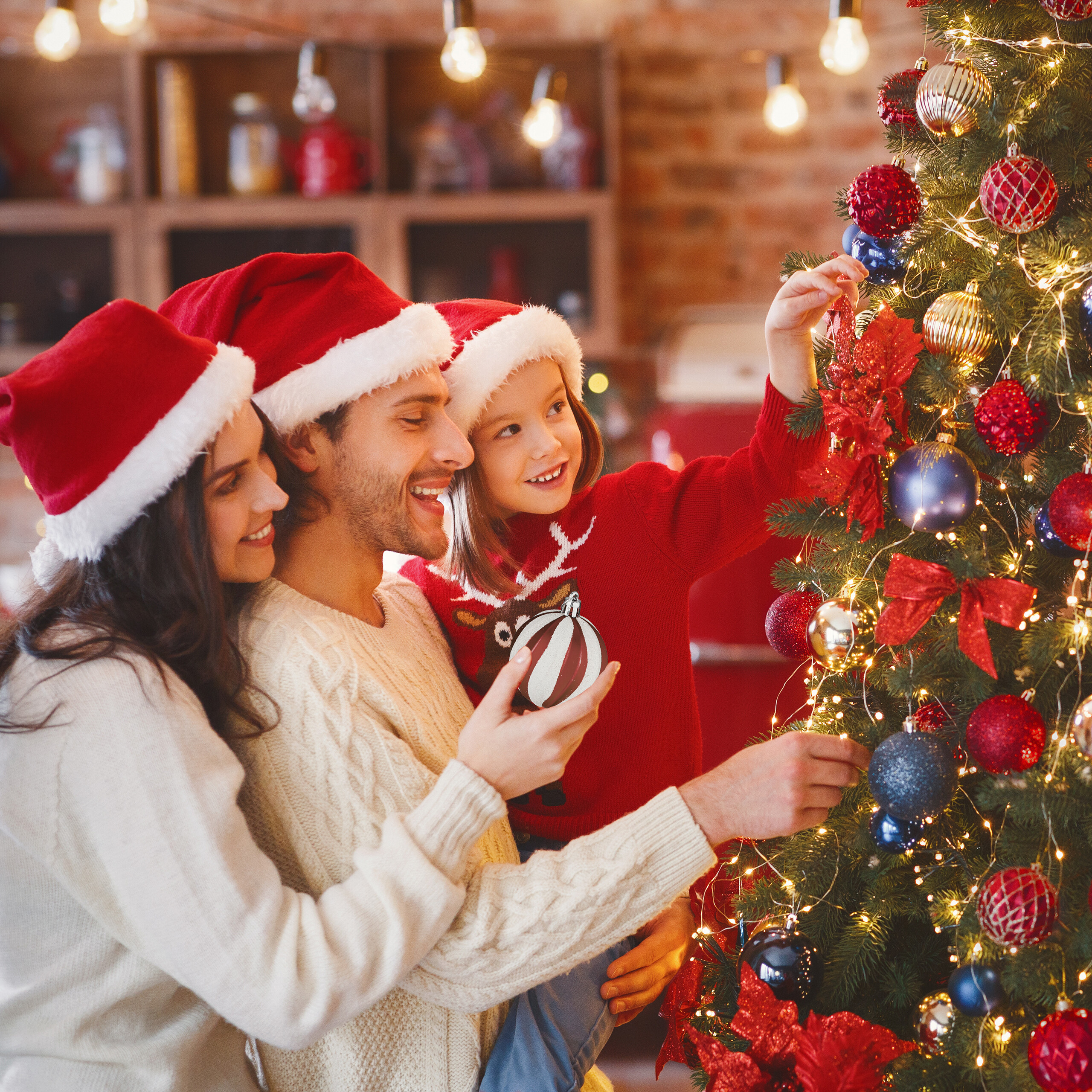 Eine lächelnde Familie in Weihnachtsmannmützen schmückt zusammen einen Weihnachtsbaum.