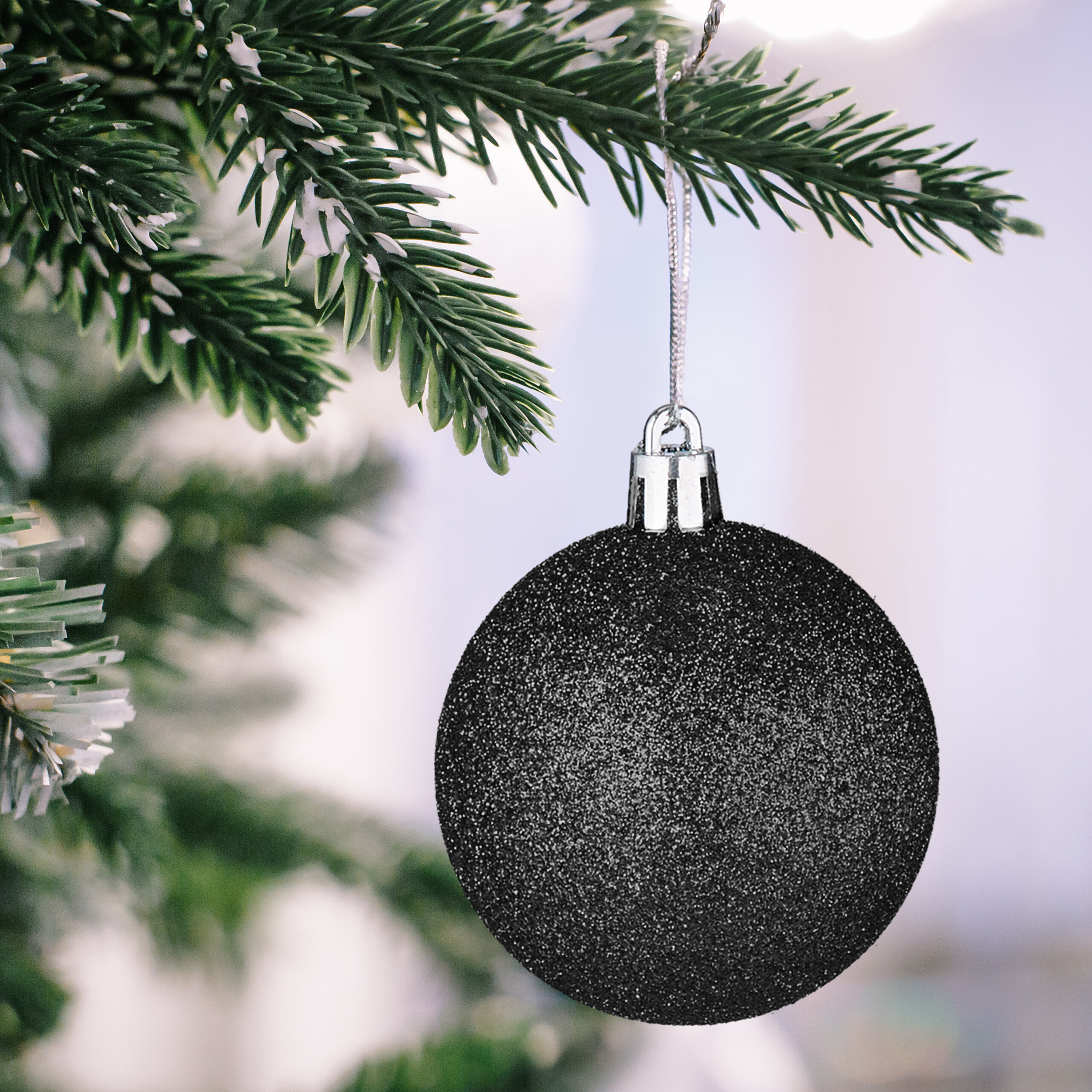 Eine glitzernde schwarze Weihnachtskugel hängt an einem verschneiten Weihnachtsbaumzweig.