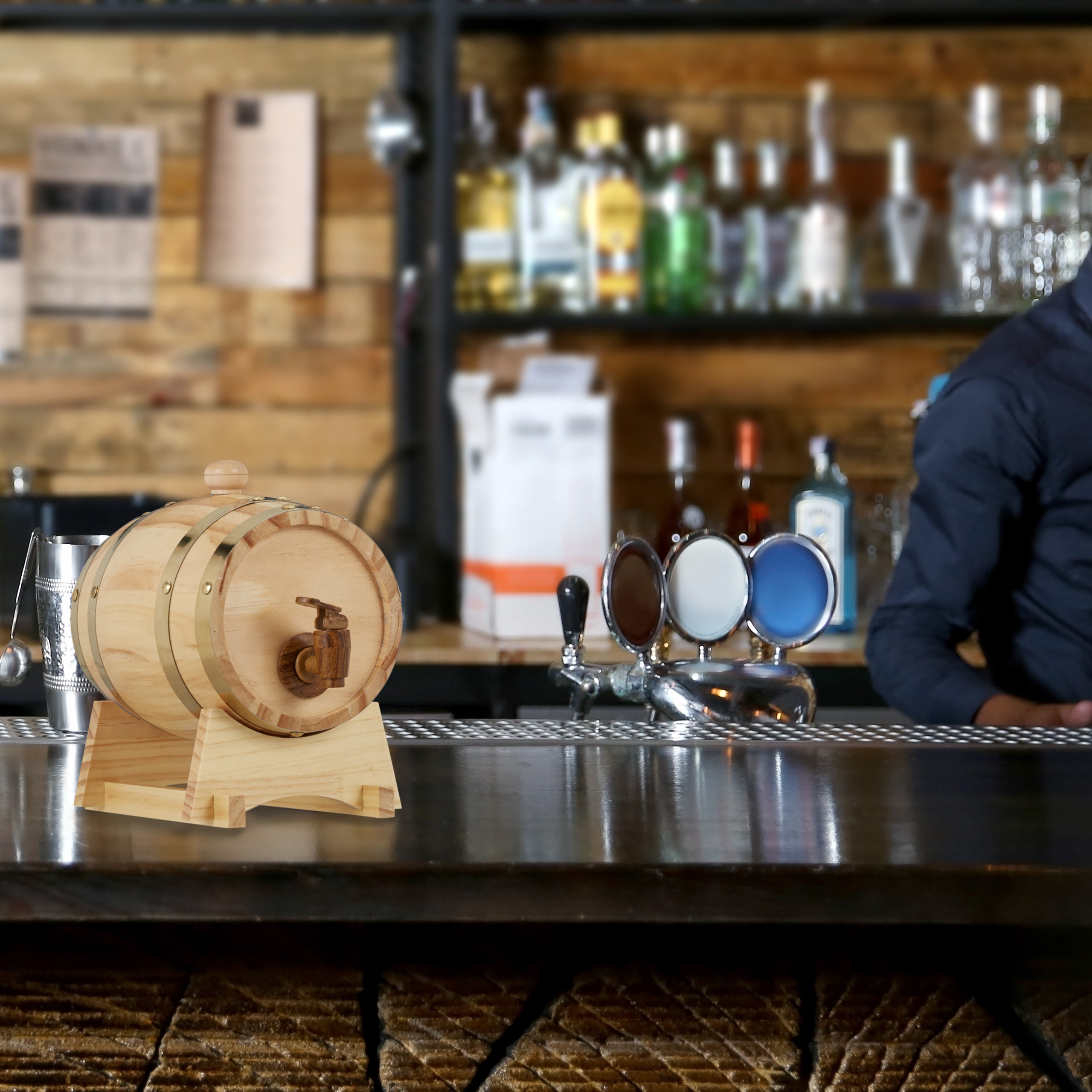 Ein kleines Holzfass mit Hahn steht auf einer Bar, im Hintergrund sind Flaschen und ein Bartender zu sehen.