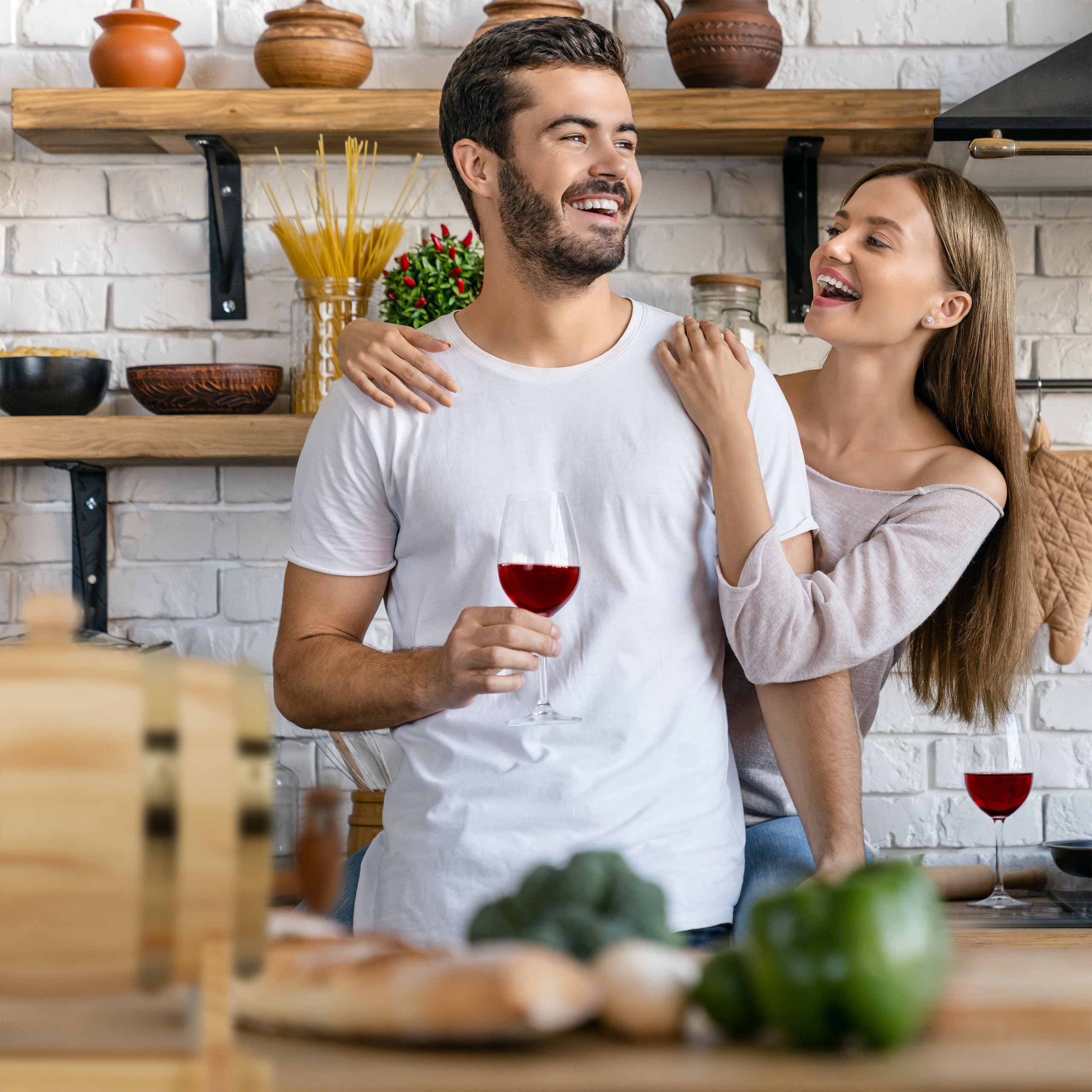 Glückliches Paar in einer Küche; der Mann hält ein Glas Rotwein, die Frau lächelt und umarmt ihn von hinten.
