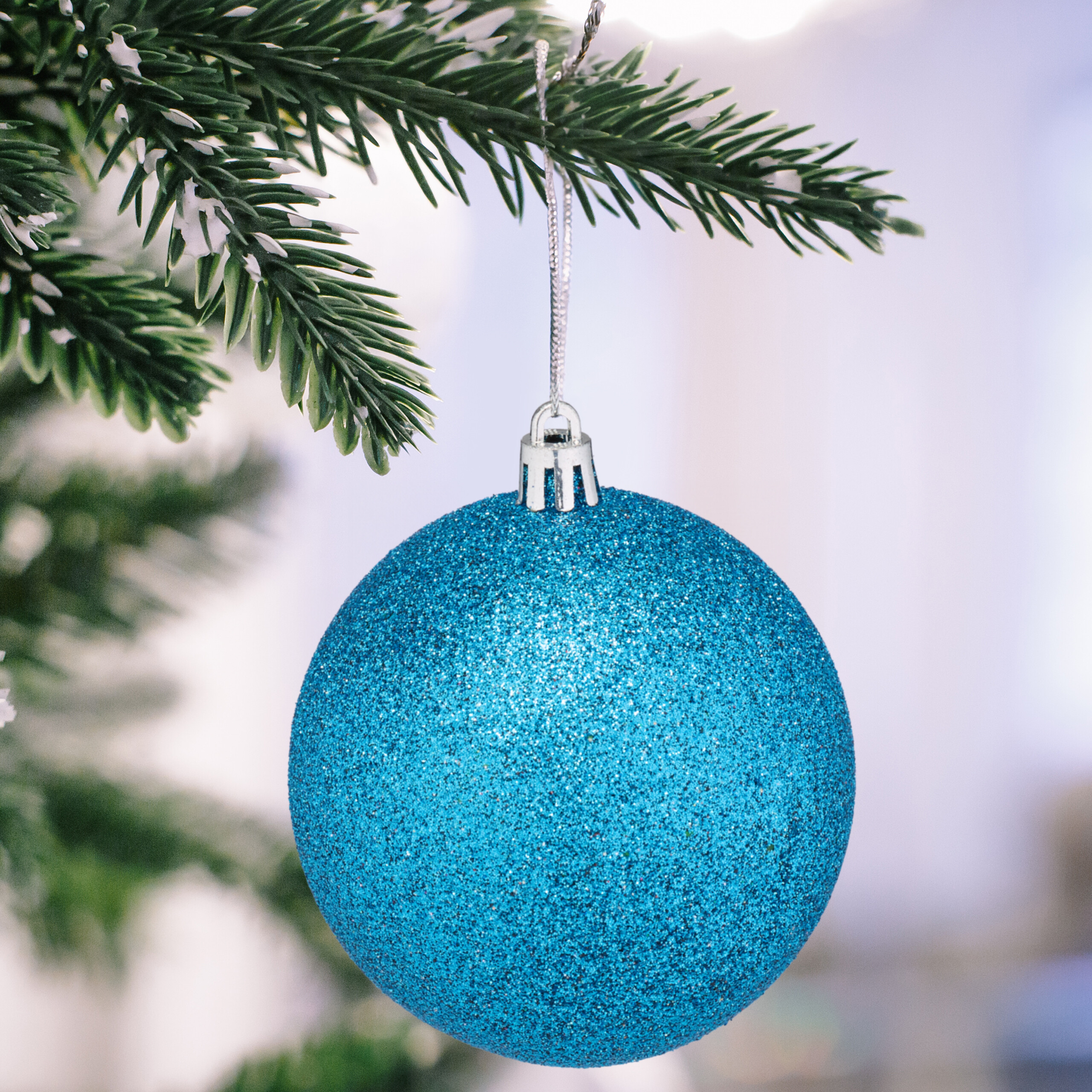 Eine glitzernde blaue Weihnachtskugel hängt an einem verschneiten Tannenzweig.