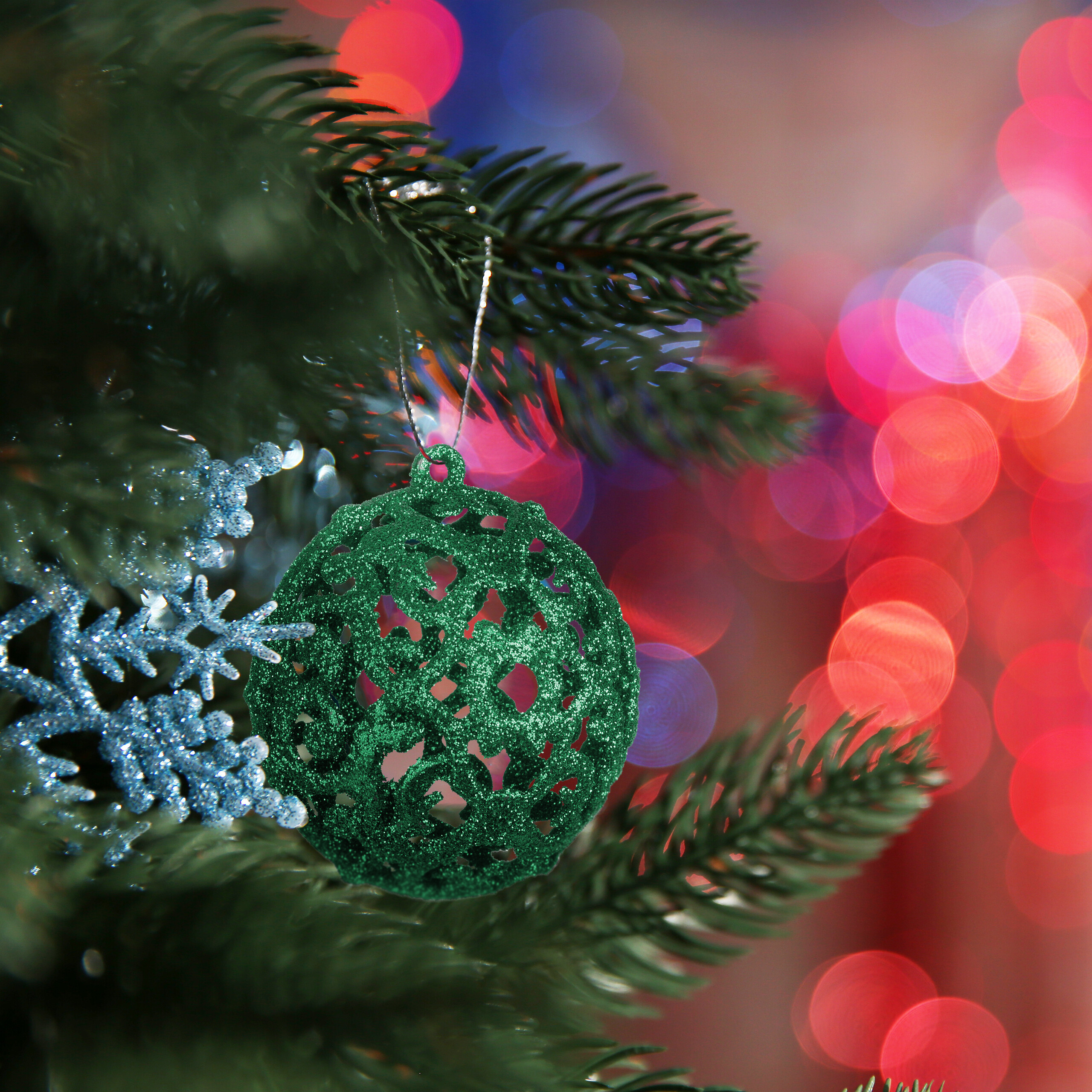 Glitzernde grüne Weihnachtskugel und silberne Schneeflocke am Baum vor verschwommenem, buntem Hintergrund.