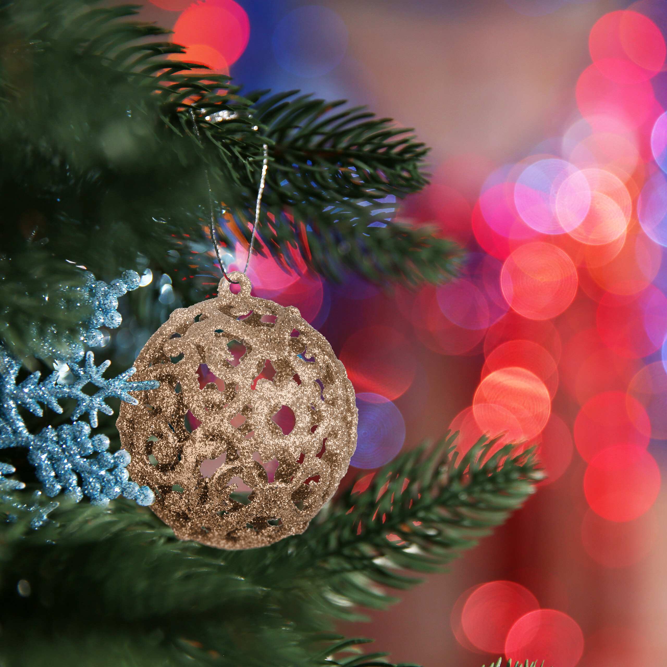 Glitzernde goldene Weihnachtskugel am Weihnachtsbaum mit blauem Schneeflockenschmuck und farbigem Bokeh im Hintergrund.