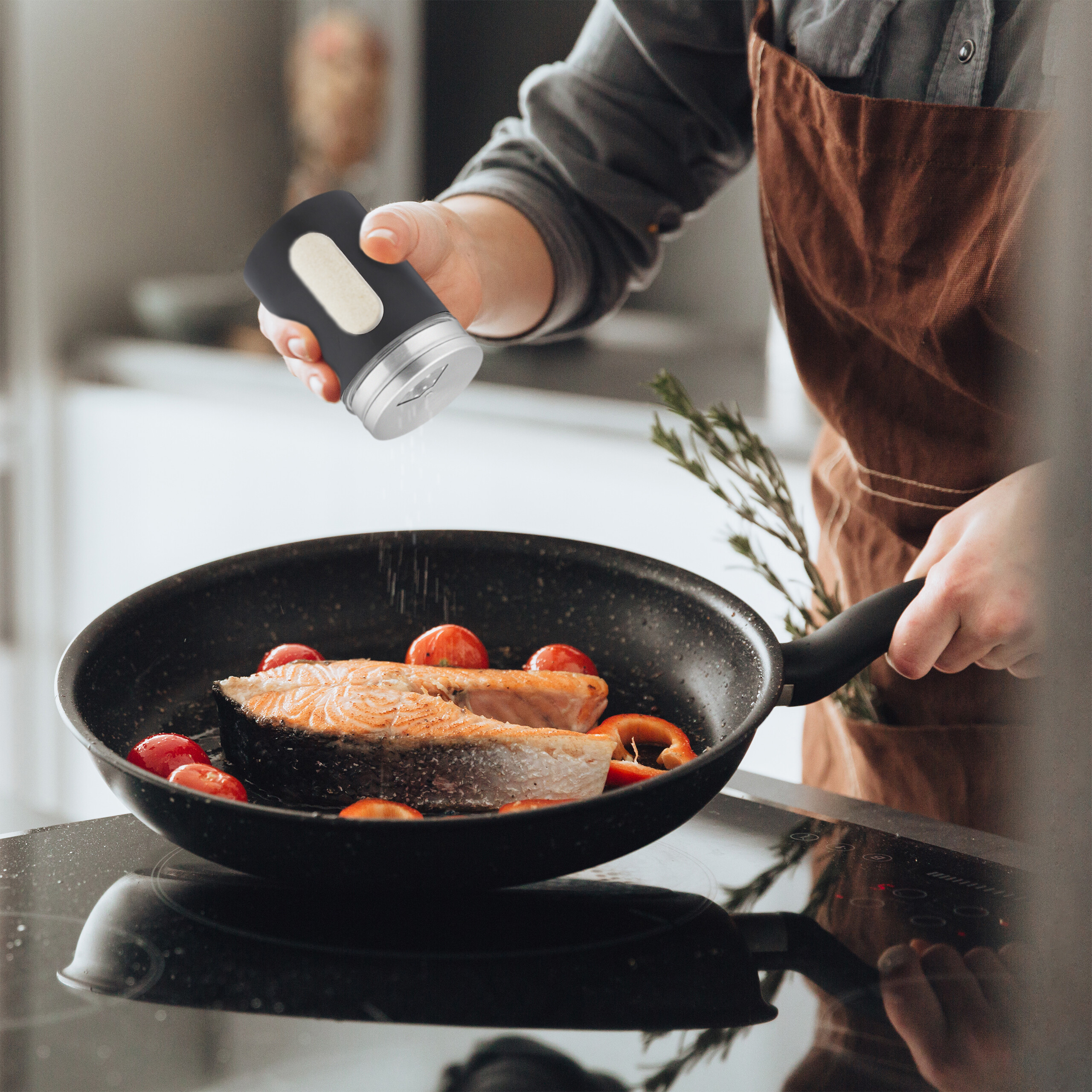 Koch streut Gewürz aus einer grauen Mühle über gebratenen Lachs und Tomaten in einer Pfanne.