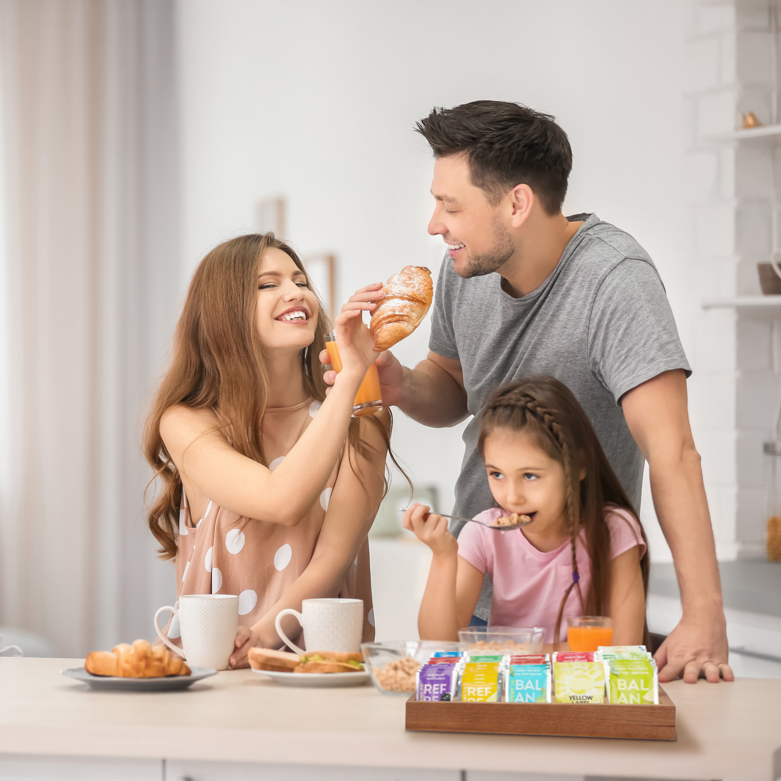 Eine glückliche Familie genießt ein Frühstück mit Croissants, Saft und Getreide in einer hellen Küche.