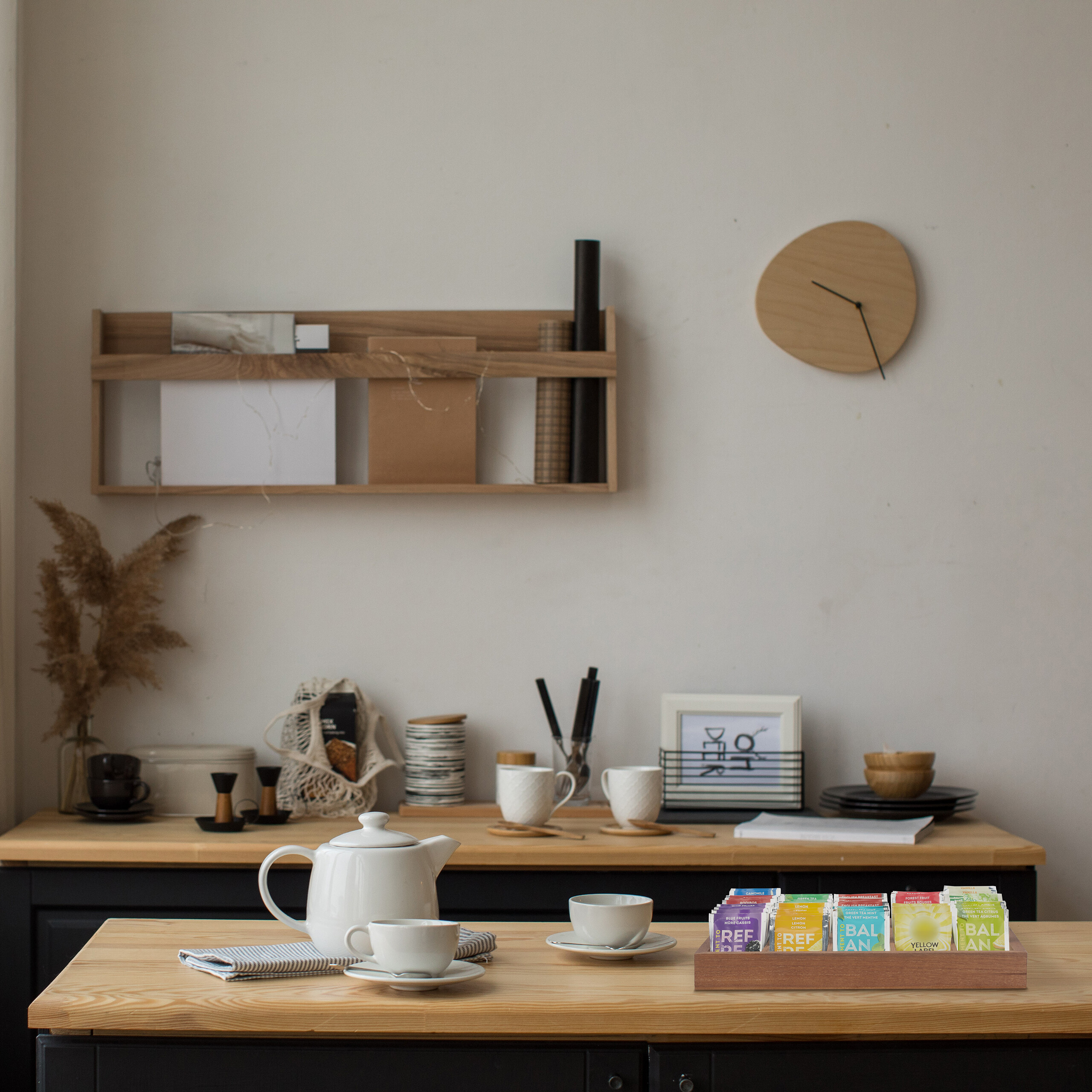 Eine weiße Teekanne und Tassen auf einem Holztisch, im Hintergrund eine Wand mit Regal und holzfarbener Uhr.