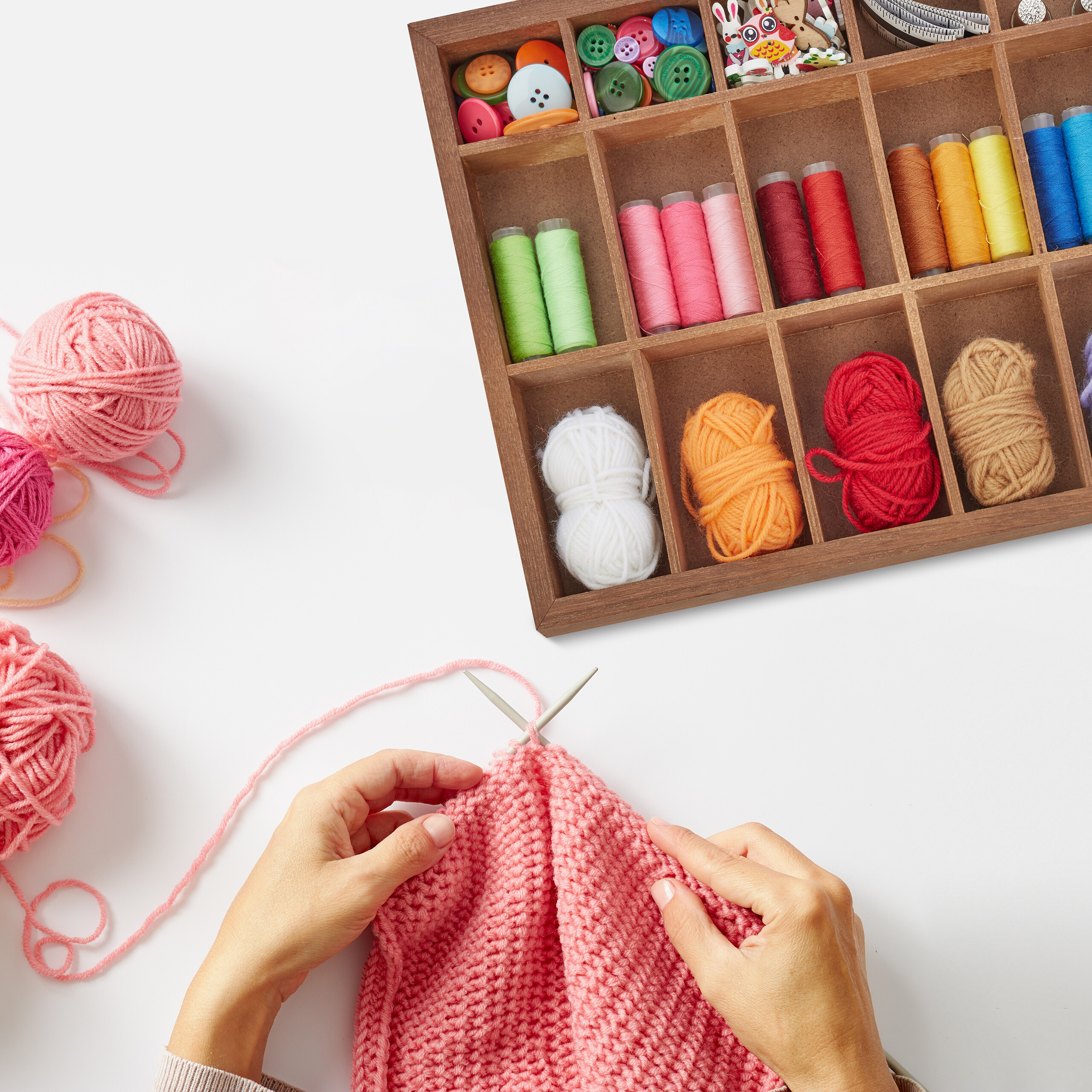 Hände stricken rosa Garn mit Nadeln, daneben eine Holzkiste voller bunter Garne und Knöpfe.