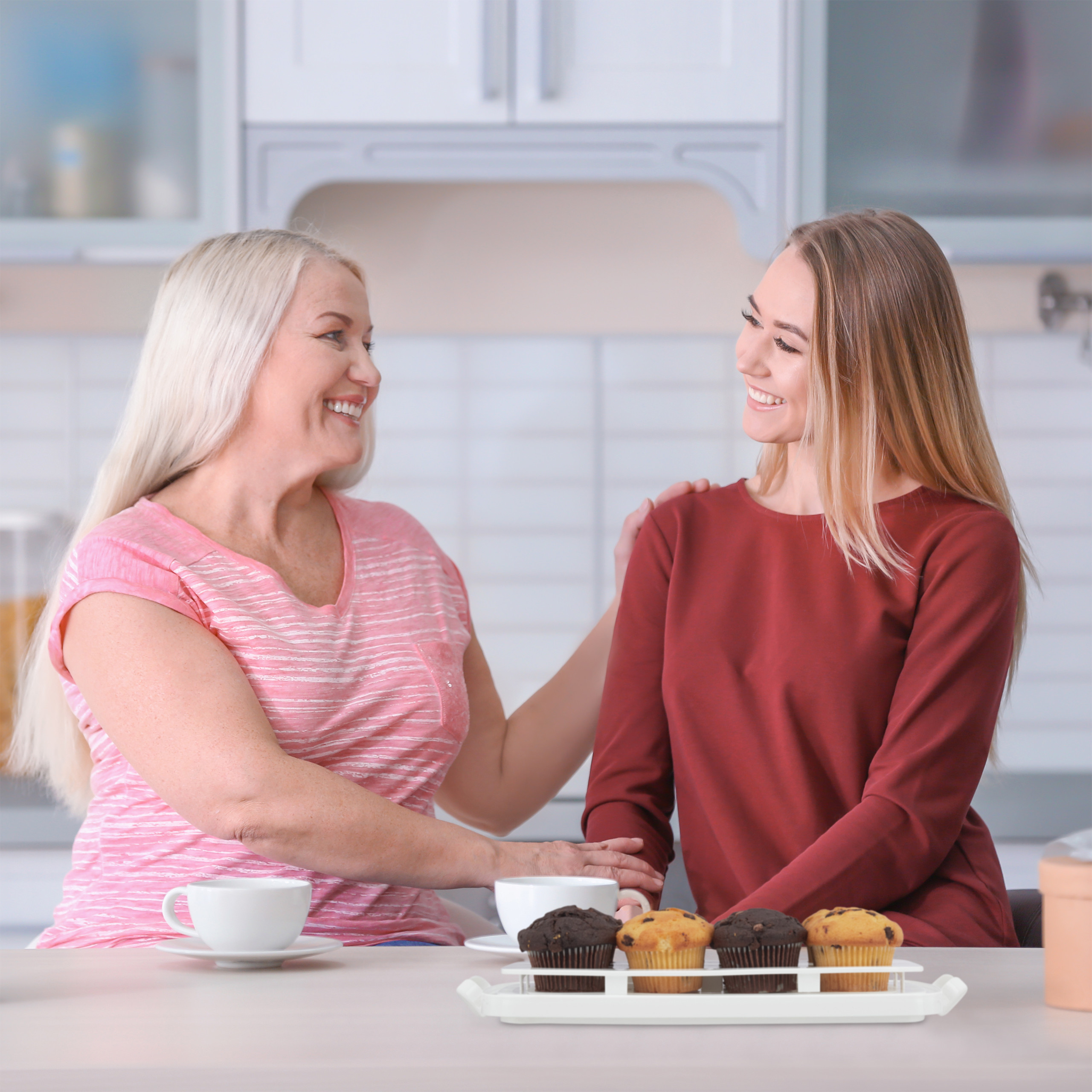 Ältere und jüngere Frau lächeln sich in einer Küche an, vor ihnen stehen Muffins und Tassen.