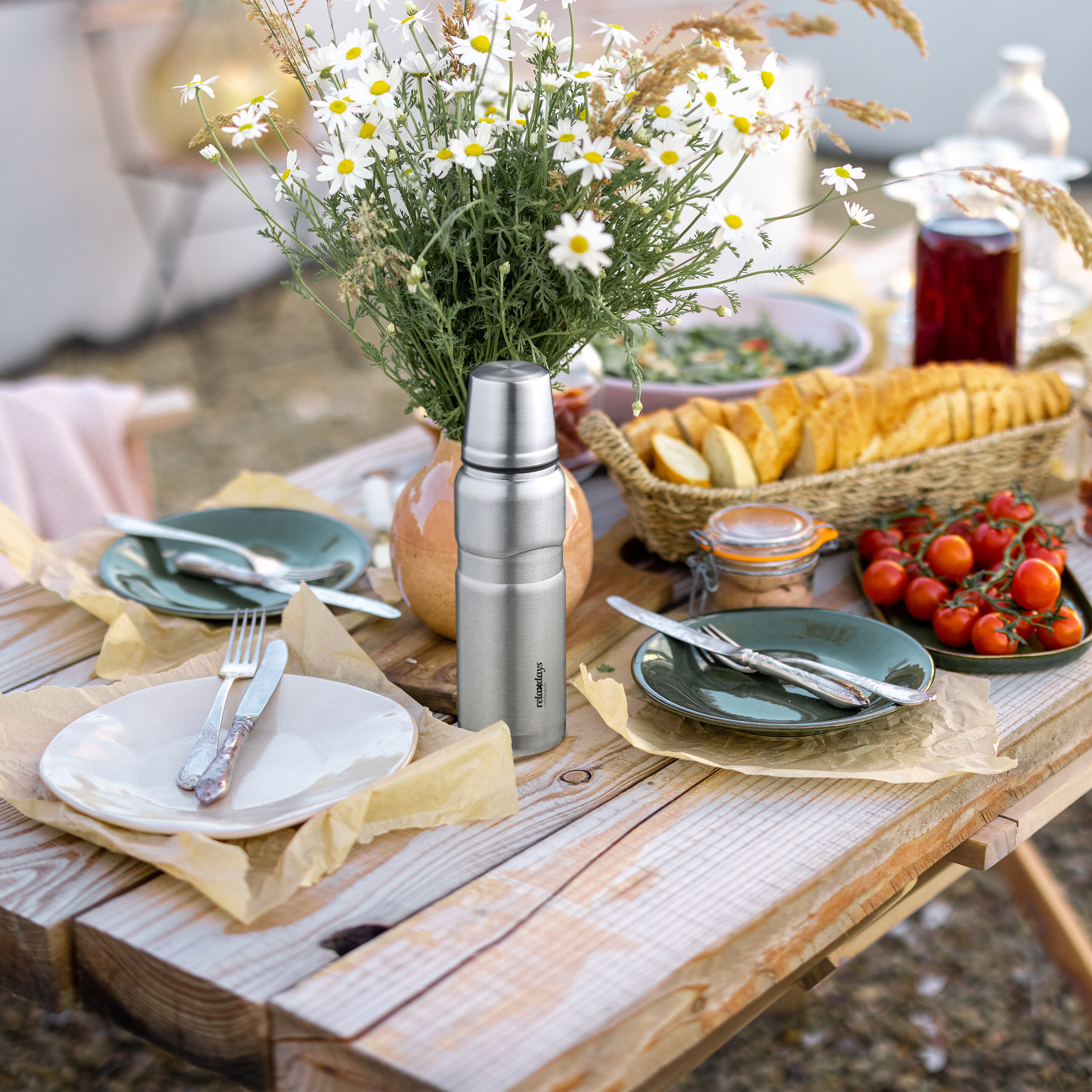 Eine silberne Thermoskanne steht auf einem Picknicktisch mit Blumen, Brot, Tellern und frischem Gemüse.